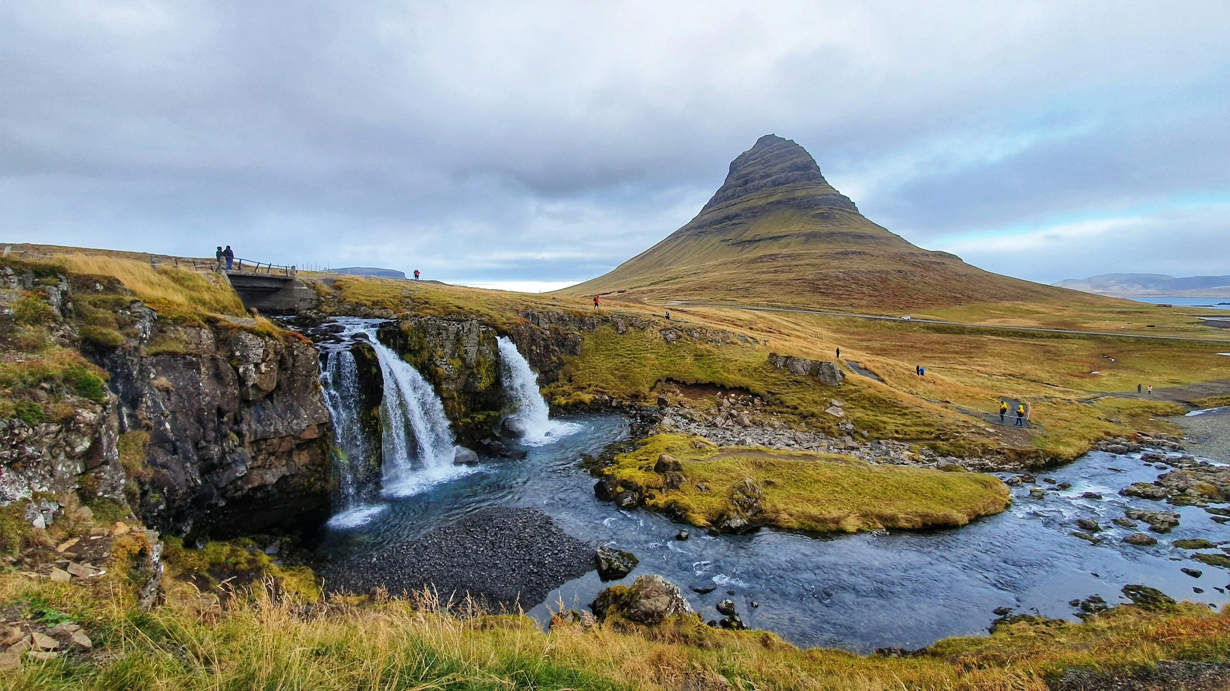Scenic landscape featuring a waterfall flowing into a river, grassy terrain, and a distinctive conical mountain in the background, with a cloudy sky and several people walking near the water.