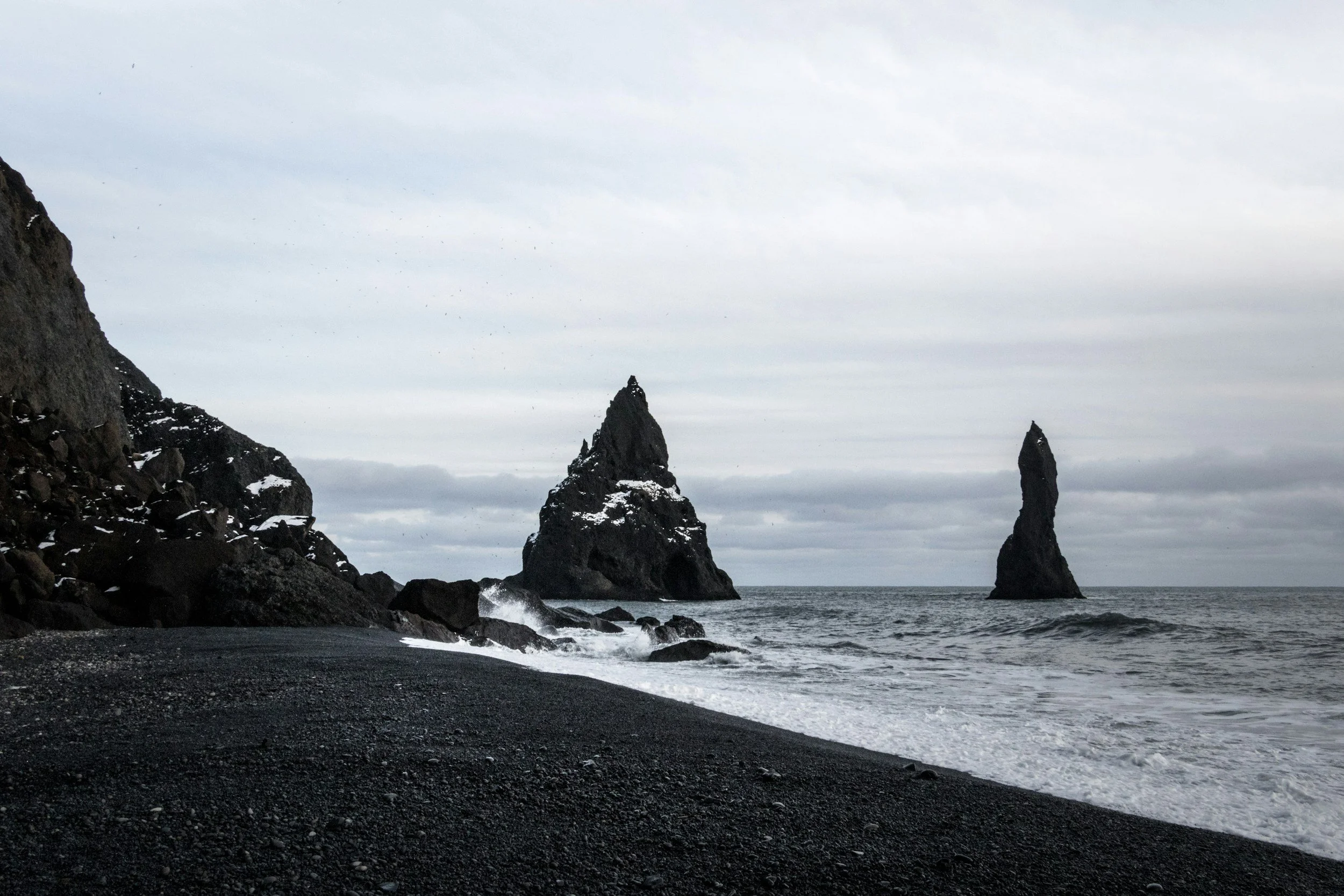A black sand beach with dark rocky cliffs on the left side, two tall jagged sea stacks in the water, and an overcast sky.