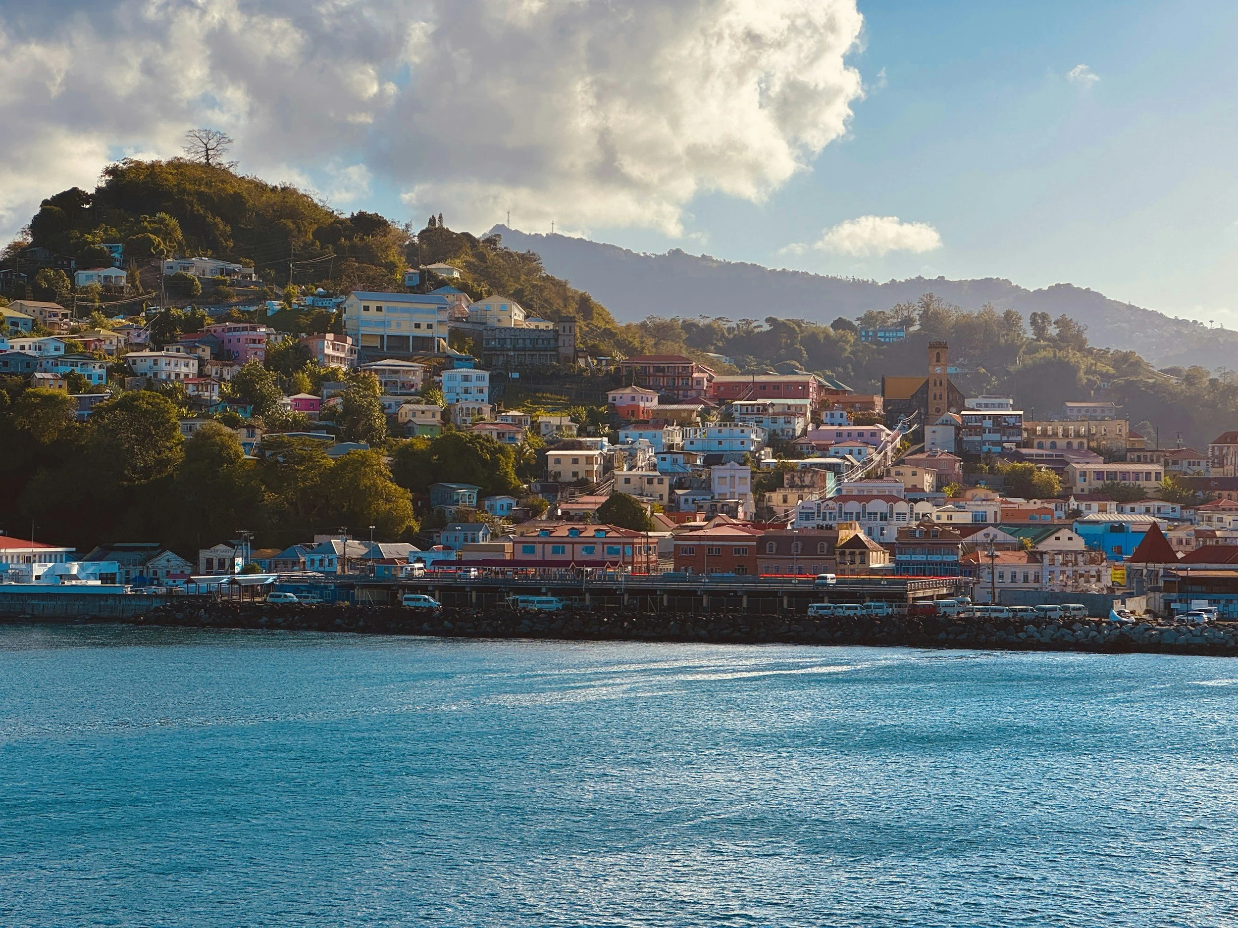Hilly coastal town with colorful houses along the shoreline and mountains in the background, under a cloudy sky.
