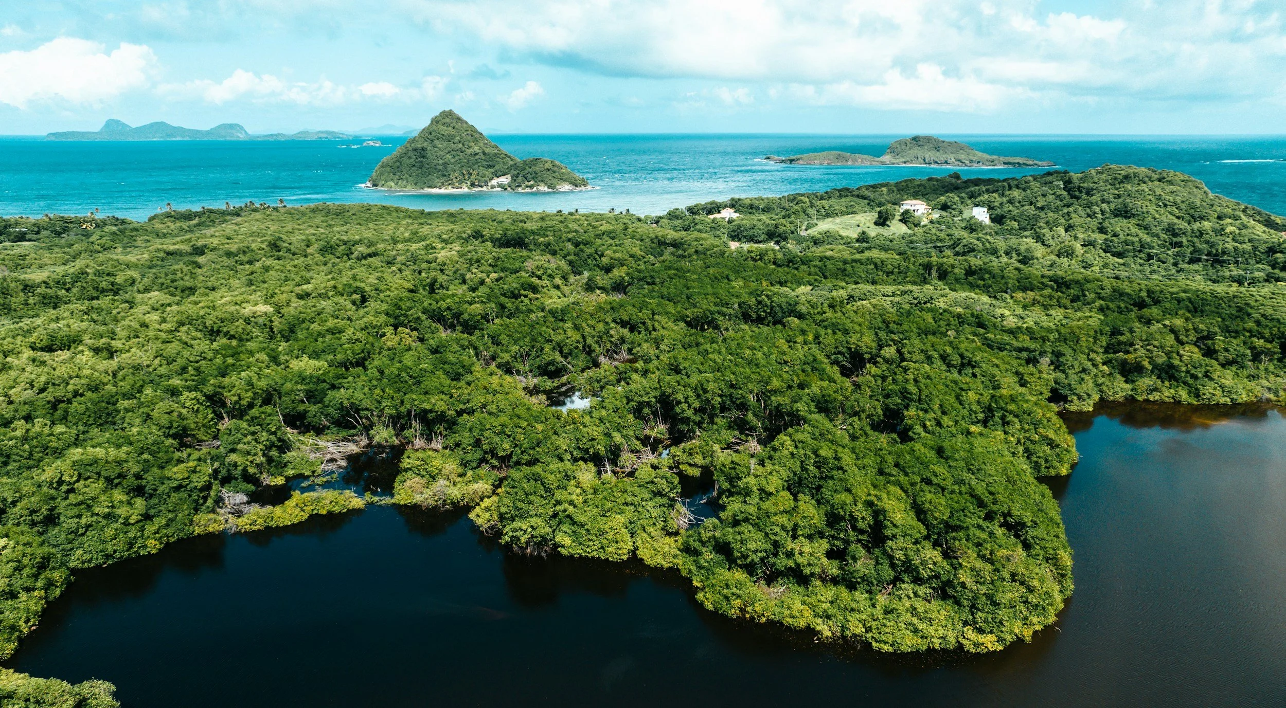 Aerial view of lush green islands and water bodies in a coastal area, with the ocean in the background and a partly cloudy sky.