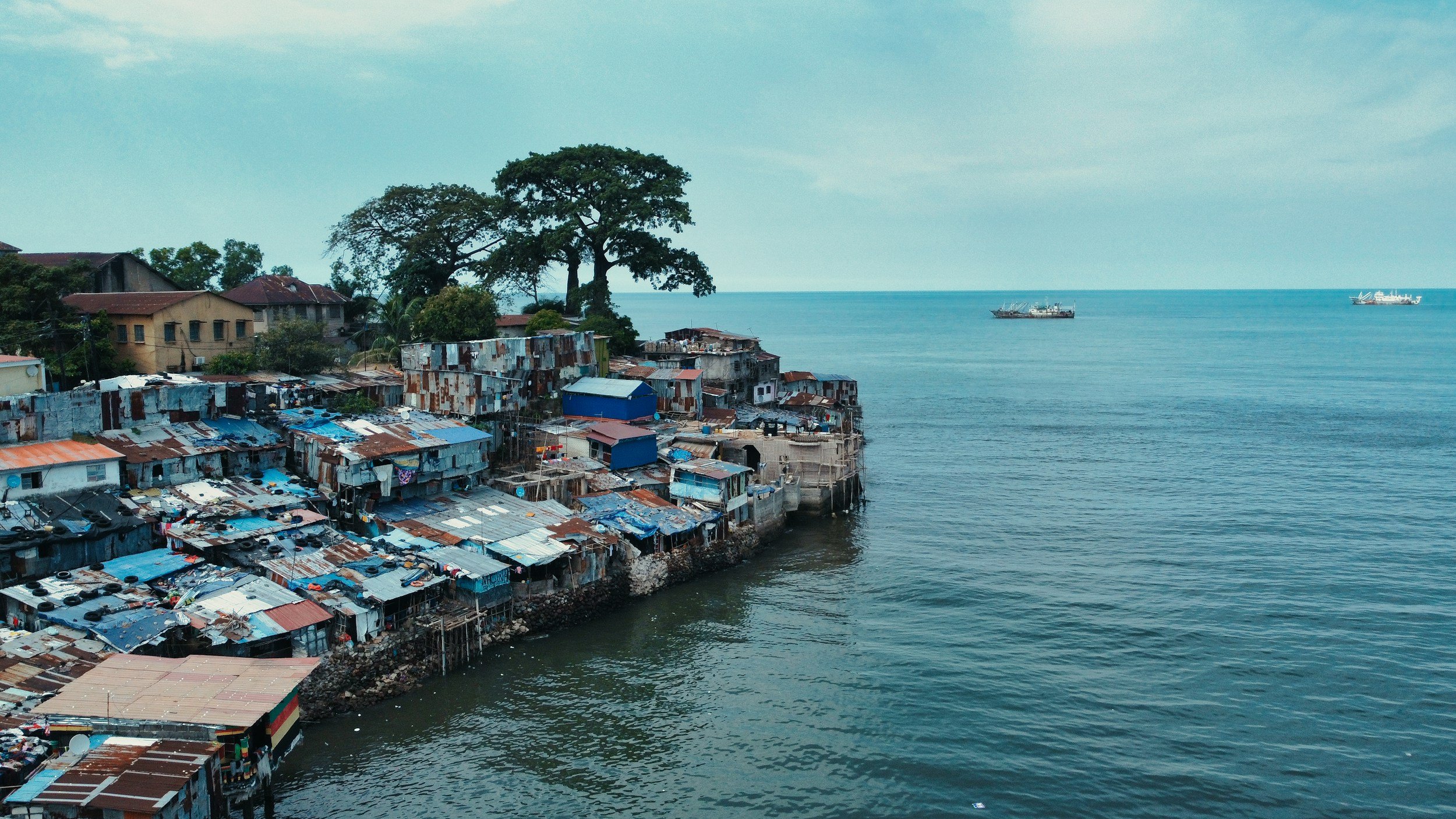 A coastal area with informal shanty houses built on stilts, made of rusted metal sheets, along a rocky shoreline. There are large trees in the background, and the ocean stretches to the horizon where two ships can be seen sailing.