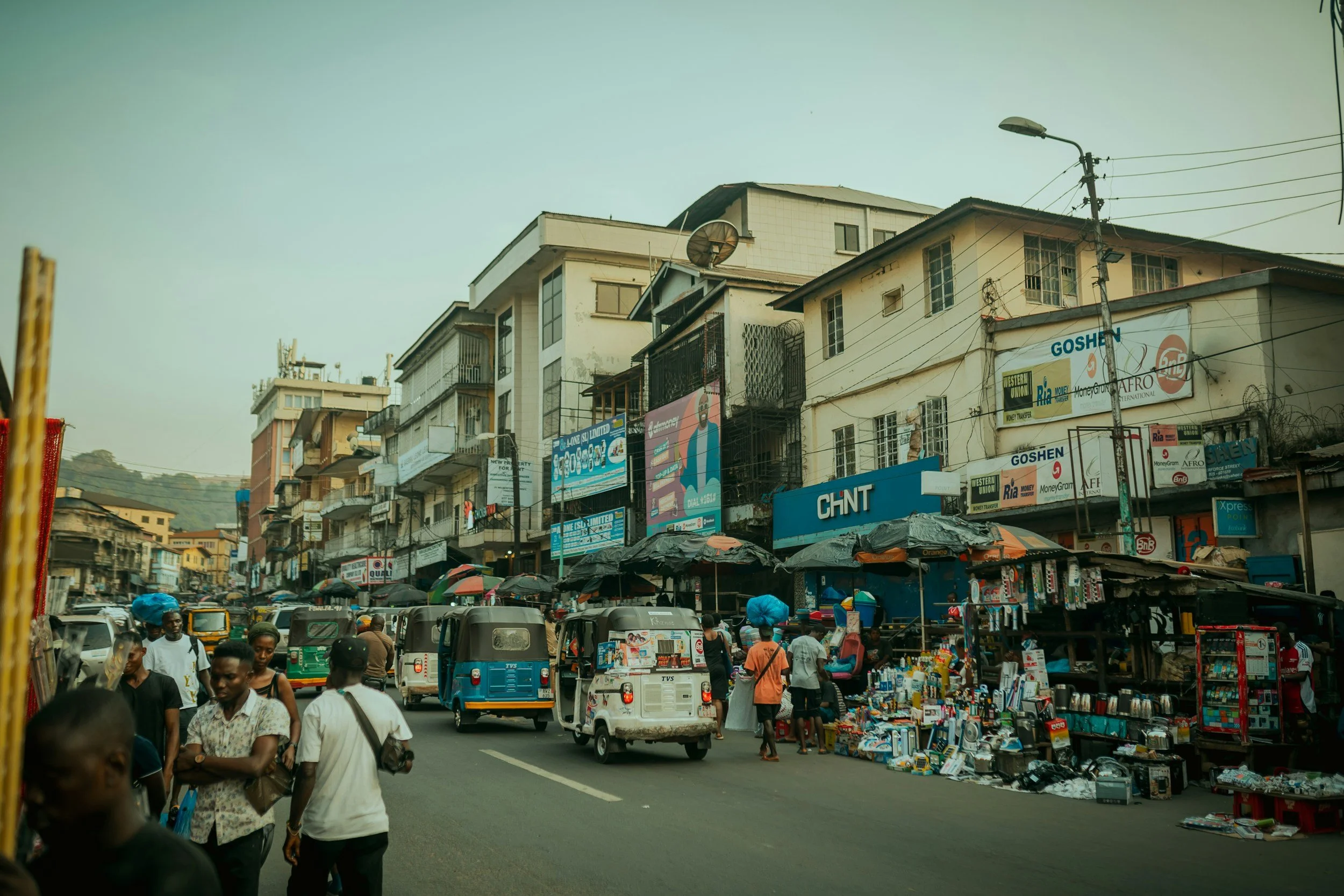 Street scene with pedestrians, auto rickshaws, market stalls, and buildings with signage in a city.