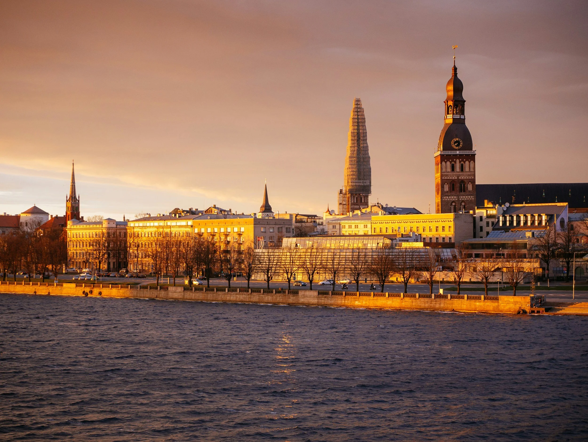 Sunset over a city skyline with historic buildings, a church with a tall clock tower, and a river in the foreground.