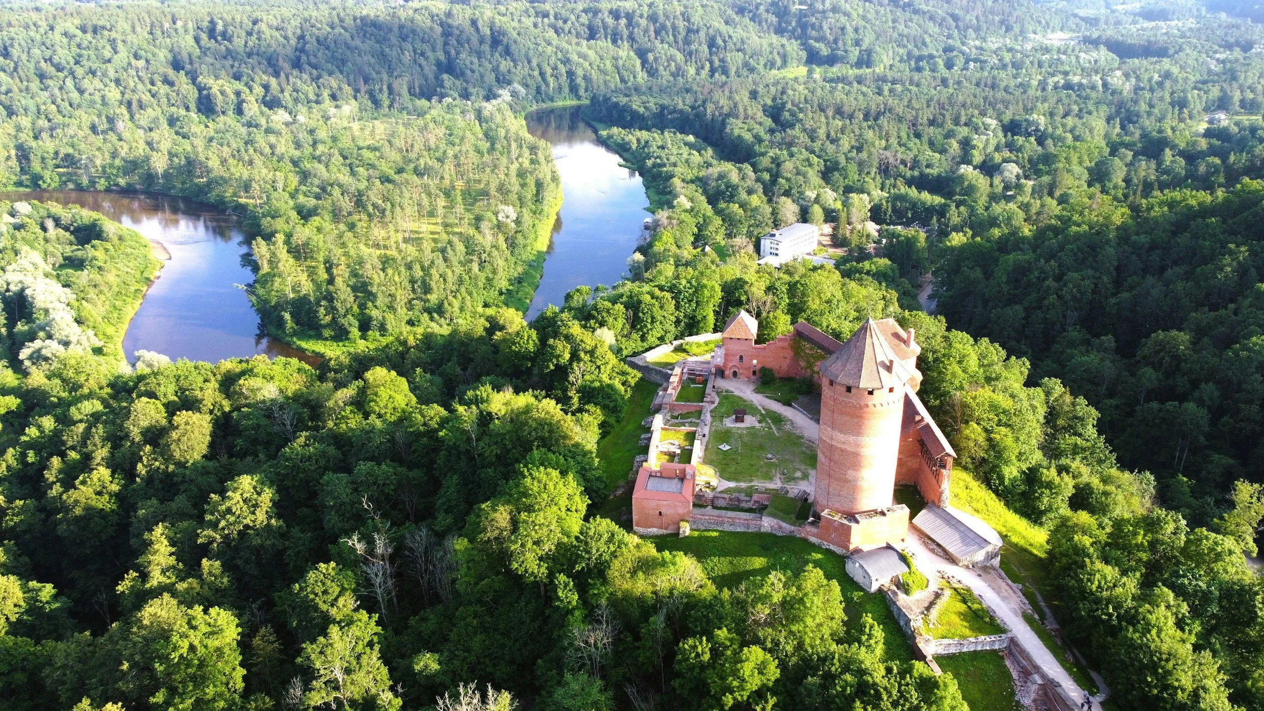 Aerial view of a historic brick castle with towers surrounded by dense green forest and a winding river in the background.