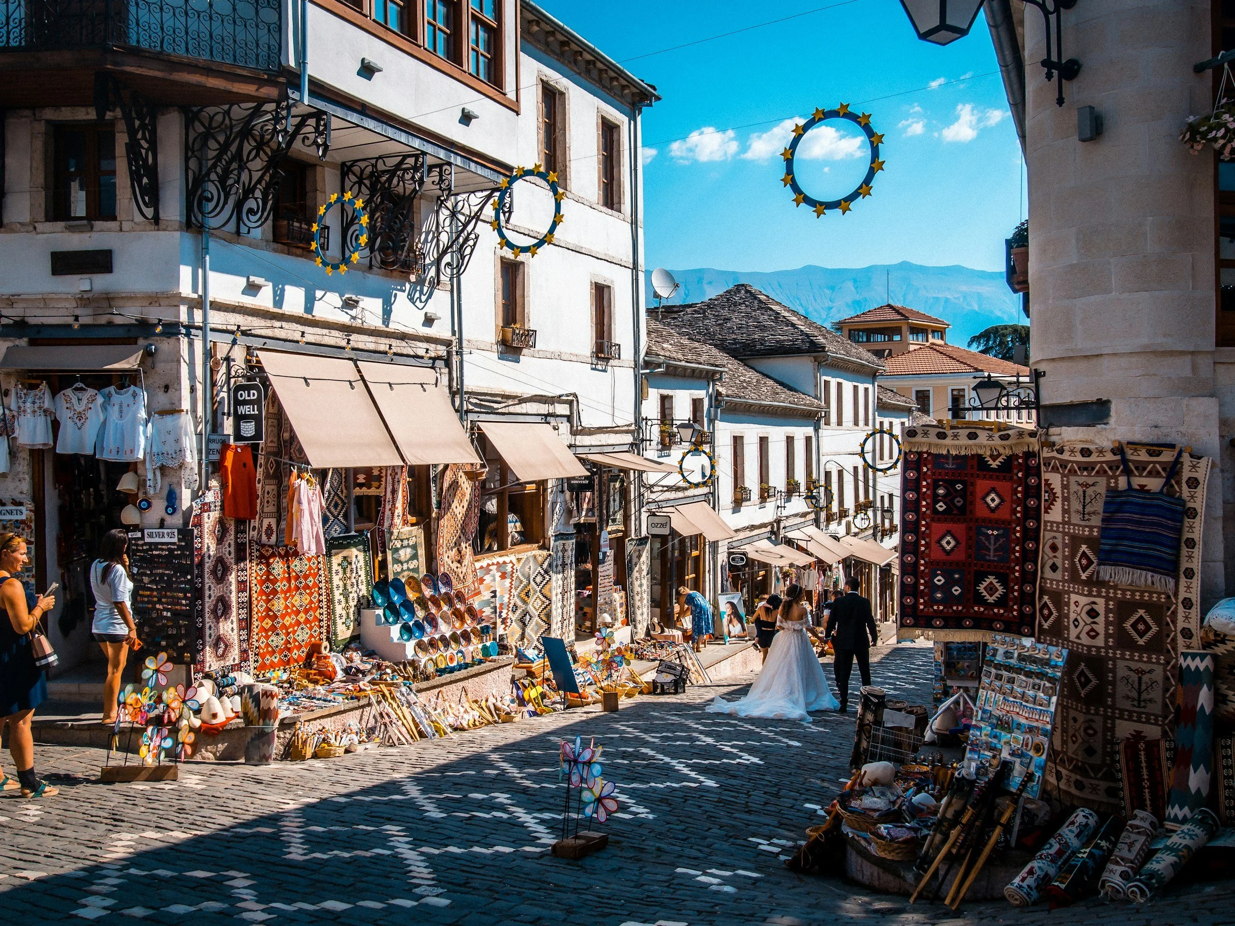 Street market with shops selling textiles, rugs, and souvenirs in a European town with mountain views in the background.