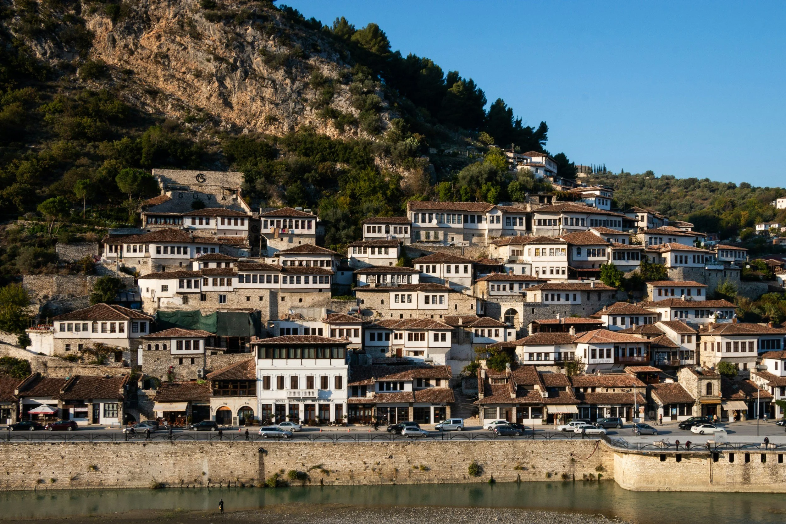 A hillside town with multiple white and gray buildings, closely packed, with some having red-tile roofs, located beside a body of water with a stone wall along the shore.