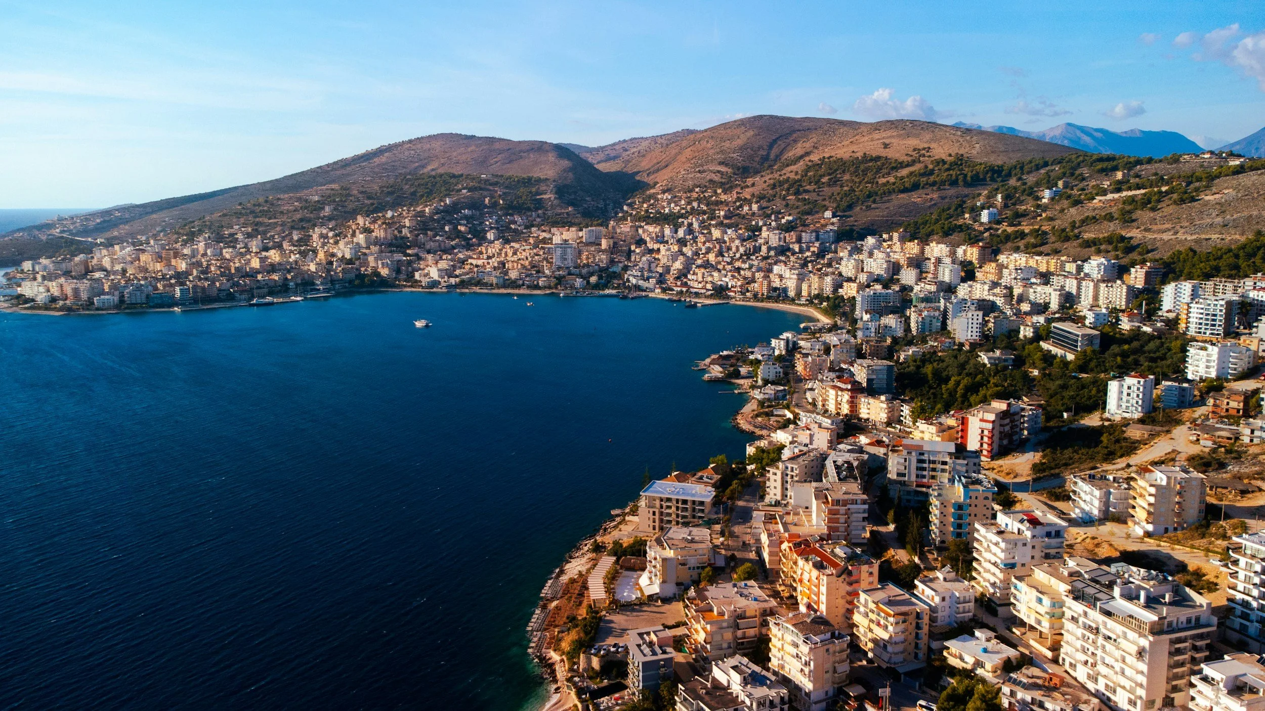 Aerial view of a coastal city with high-rise buildings along a curved shoreline, surrounded by hills and mountains in the background.