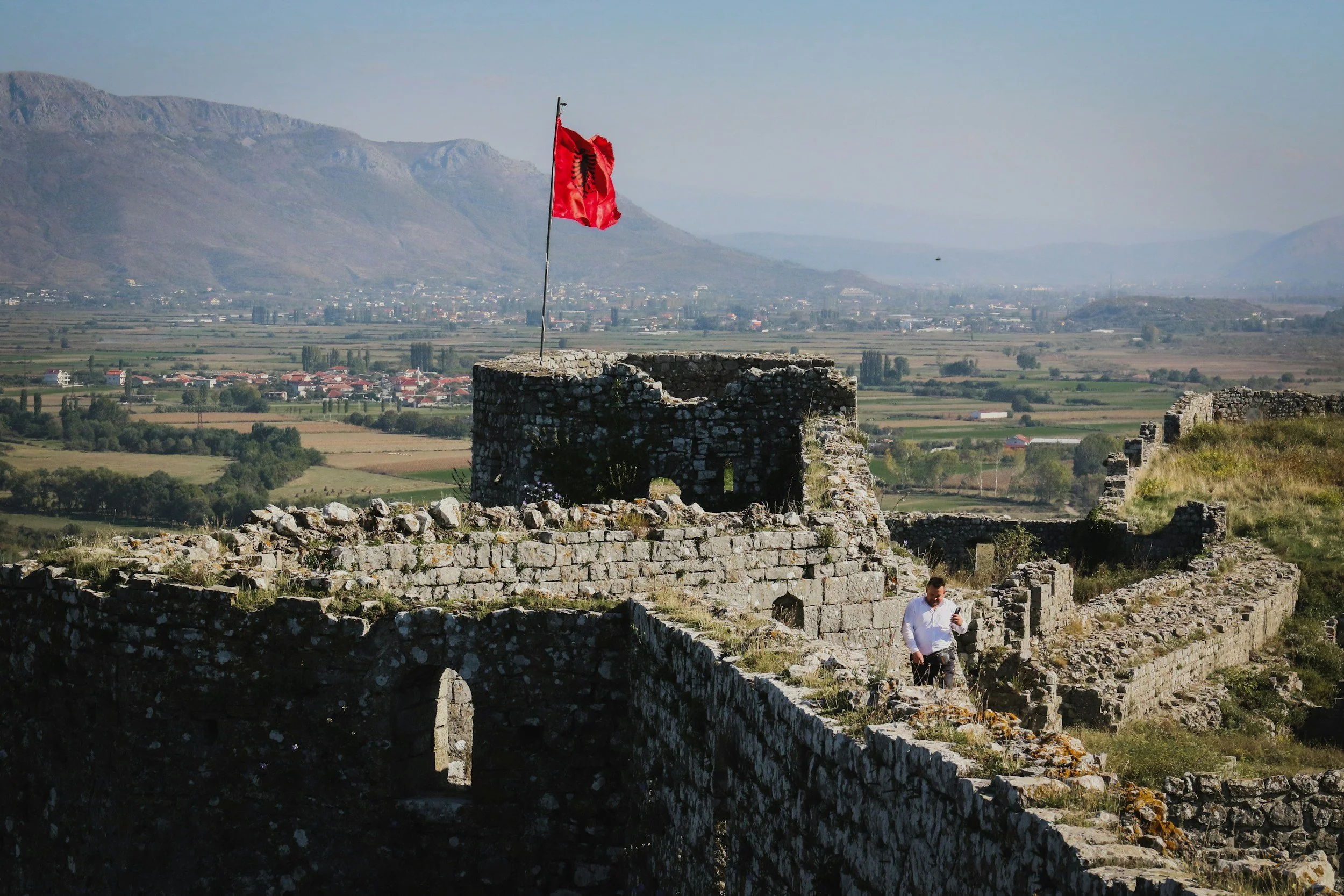 Ruins of an ancient stone castle on a hilltop, with a red flag flying on a pole. A man is walking among the ruins, holding a mobile phone, overlooking a lush green valley with mountains in the background.
