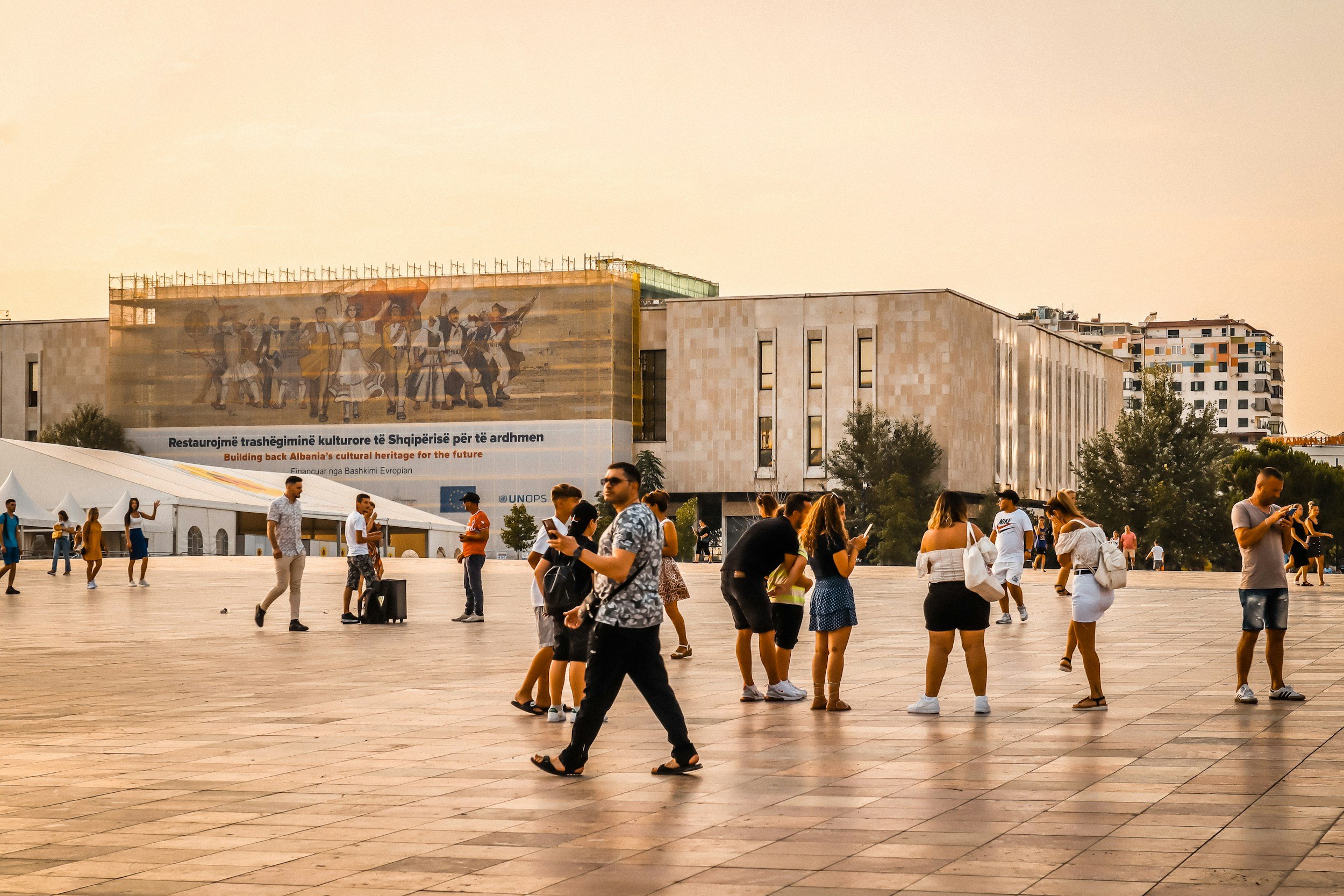 People standing and walking in an open plaza in front of a modern building with a large mural, trees, and a tent to the left. Some individuals are using their phones, and the scene is during sunset.