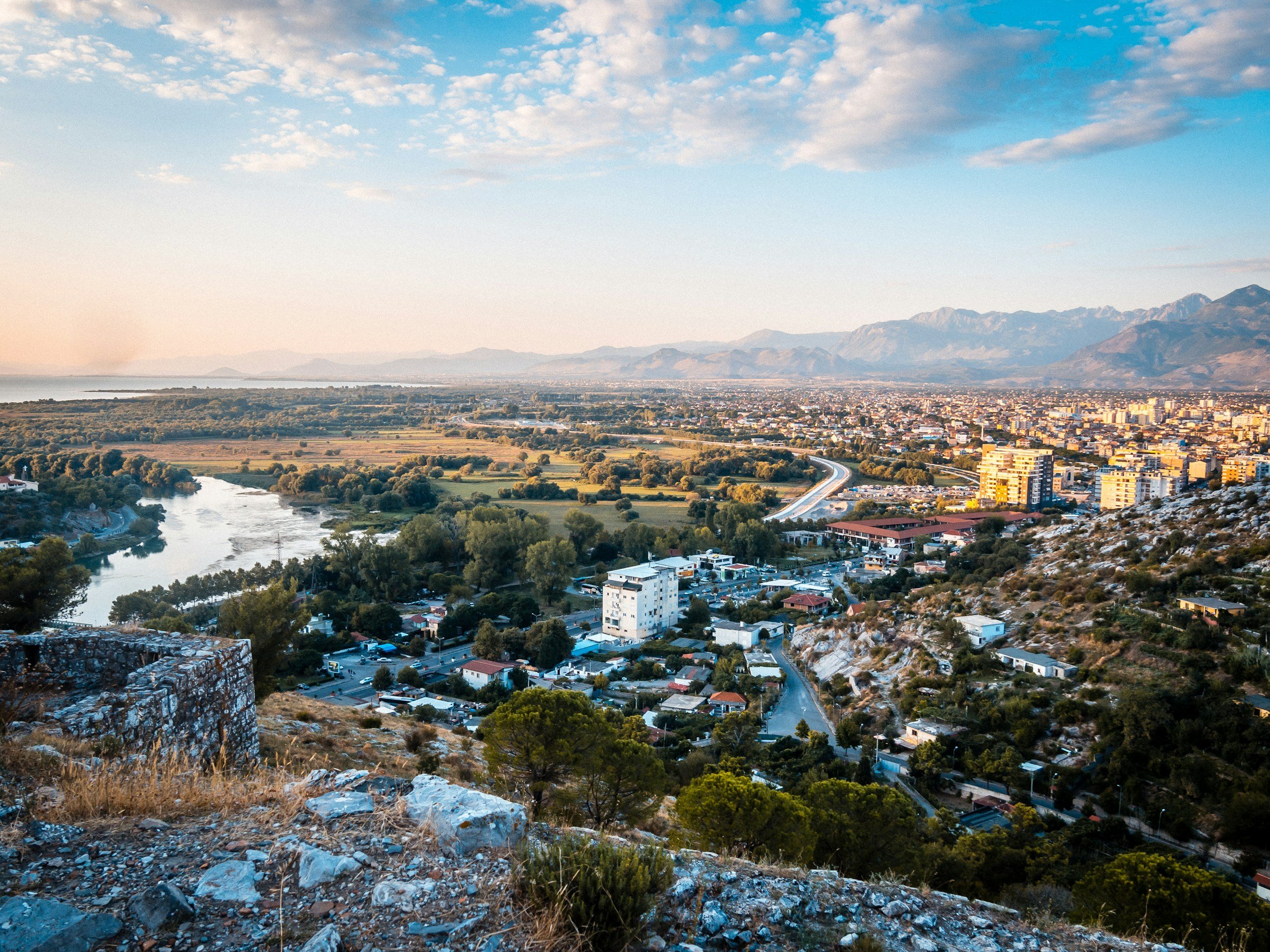 A panoramic view of a city by a river, surrounded by mountains and greenery, with a partly cloudy sky overhead.