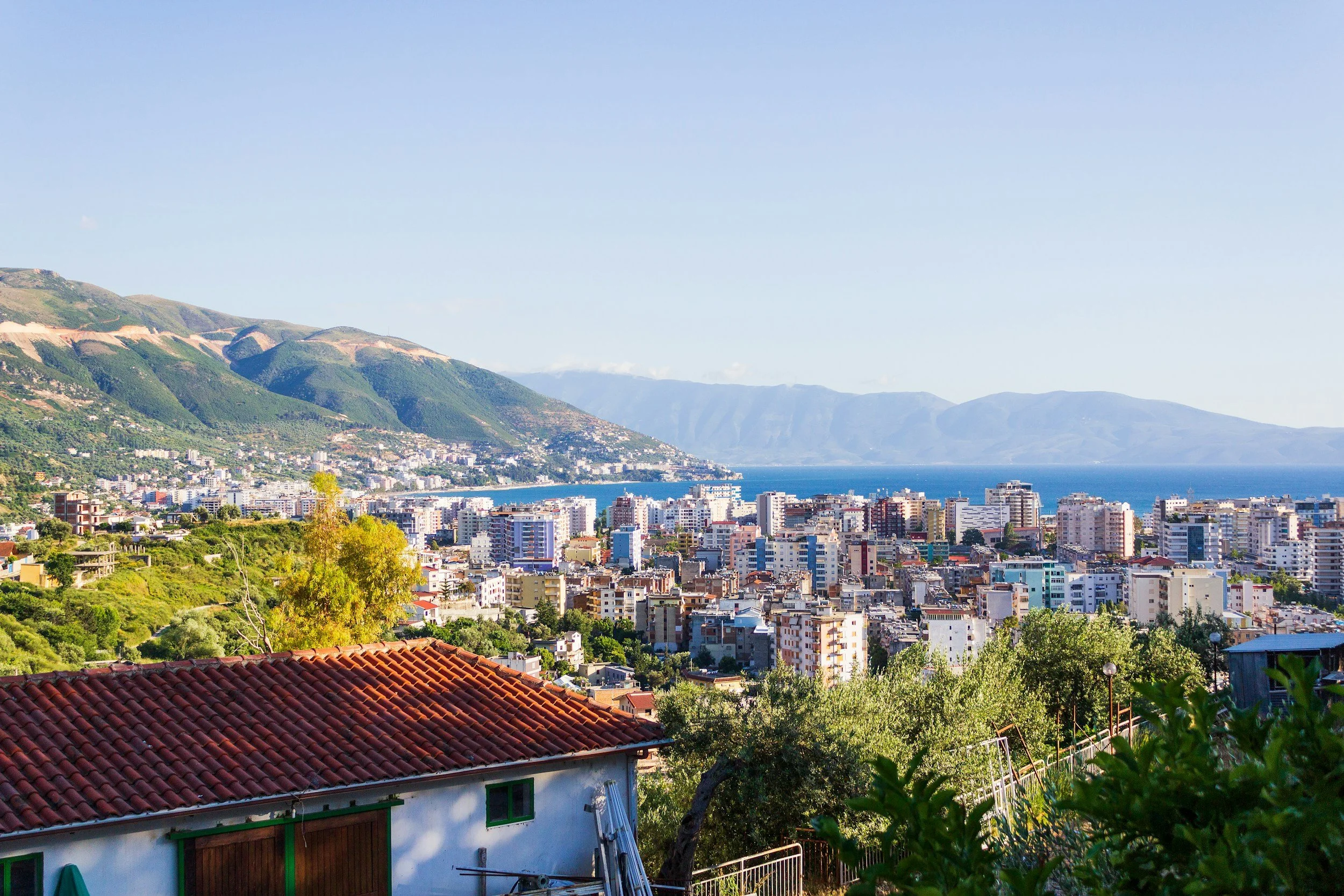 A panoramic view of a coastal city with numerous buildings, green trees, mountains in the background, and a body of water, seen from a hillside with a house that has a red-tiled roof in the foreground.