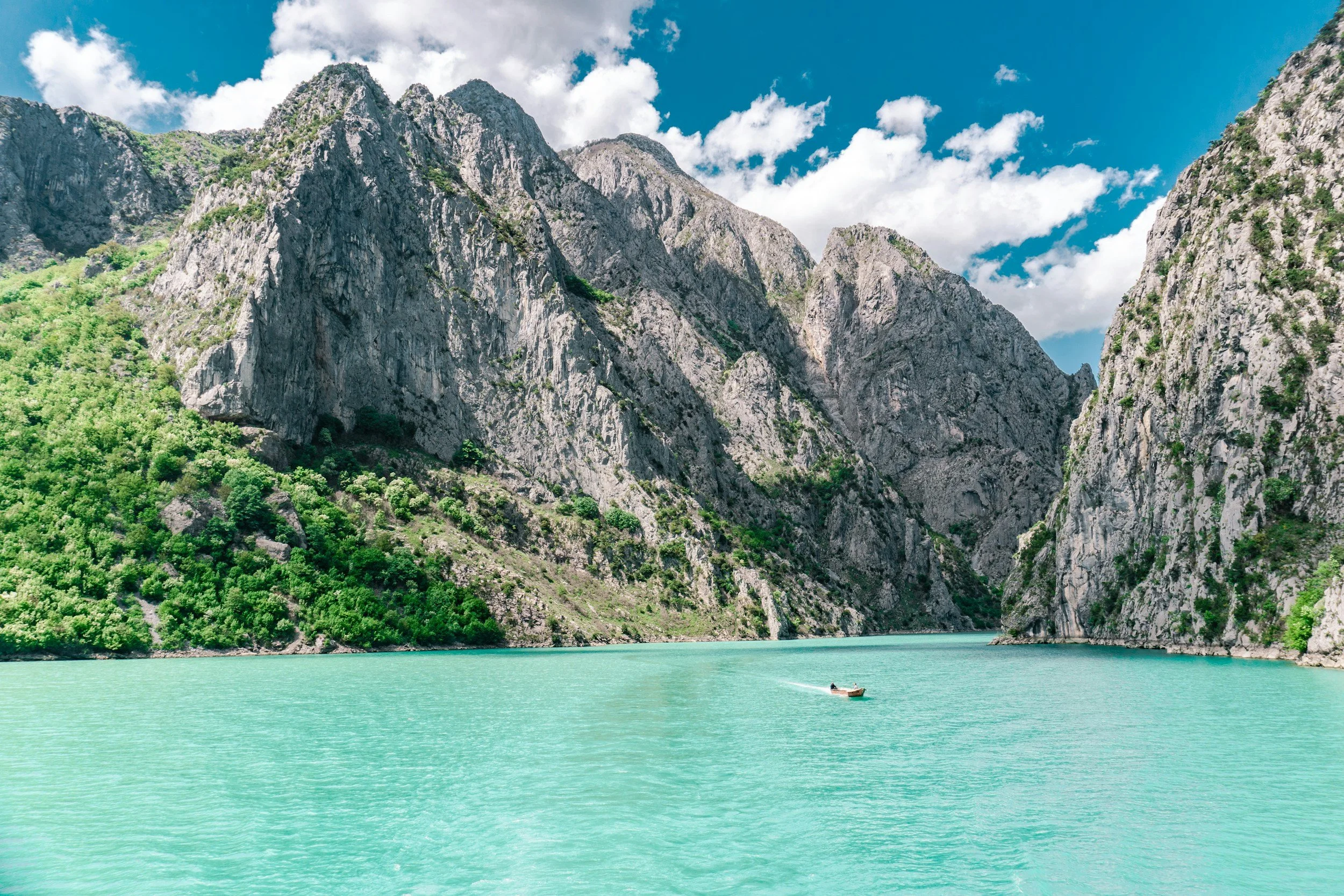 A body of turquoise water surrounded by steep rocky cliffs with patches of green vegetation, under a partly cloudy sky.
