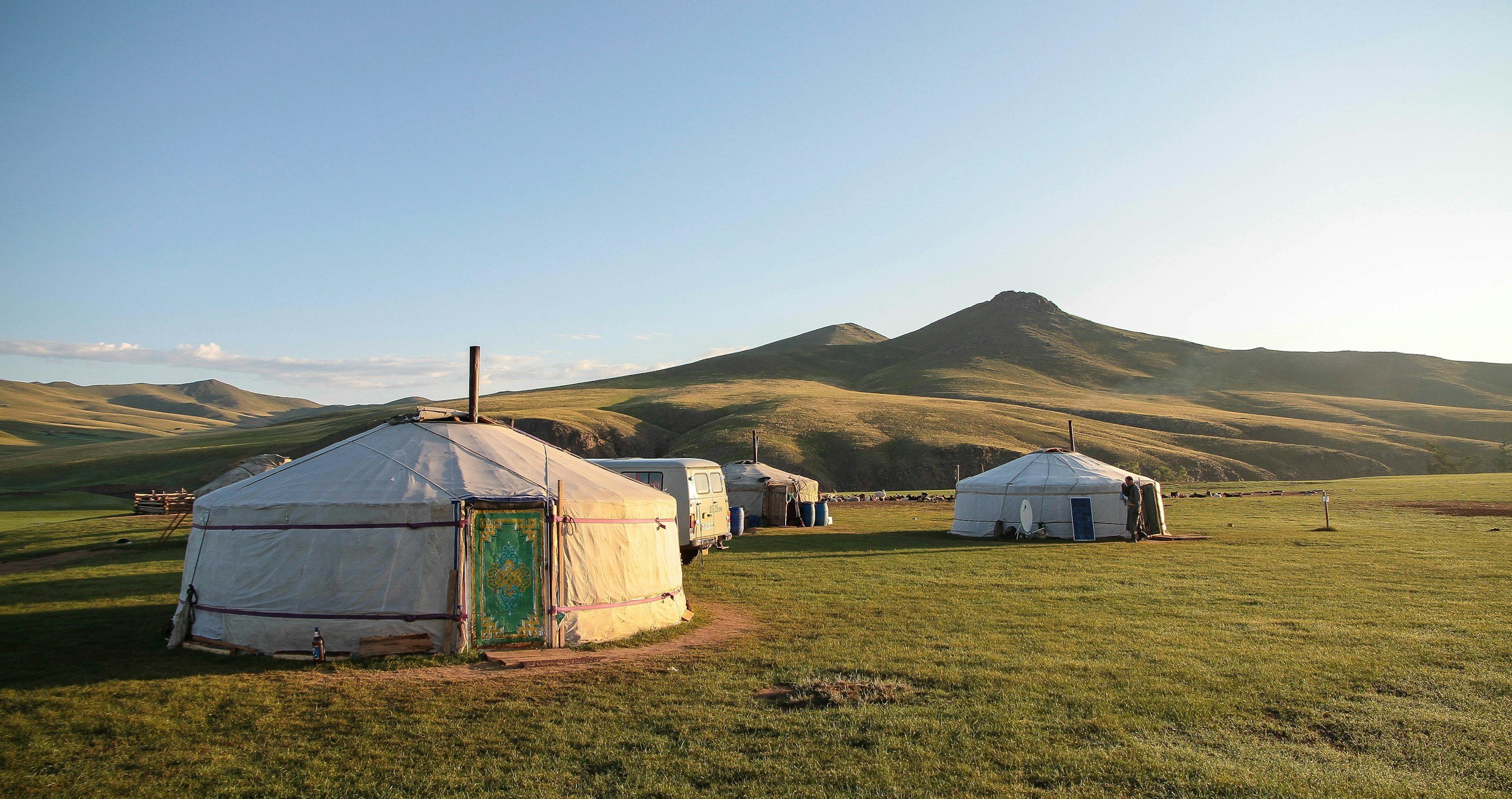 Two traditional yurts on a grassy plain with rolling hills and mountains in the background during daylight.