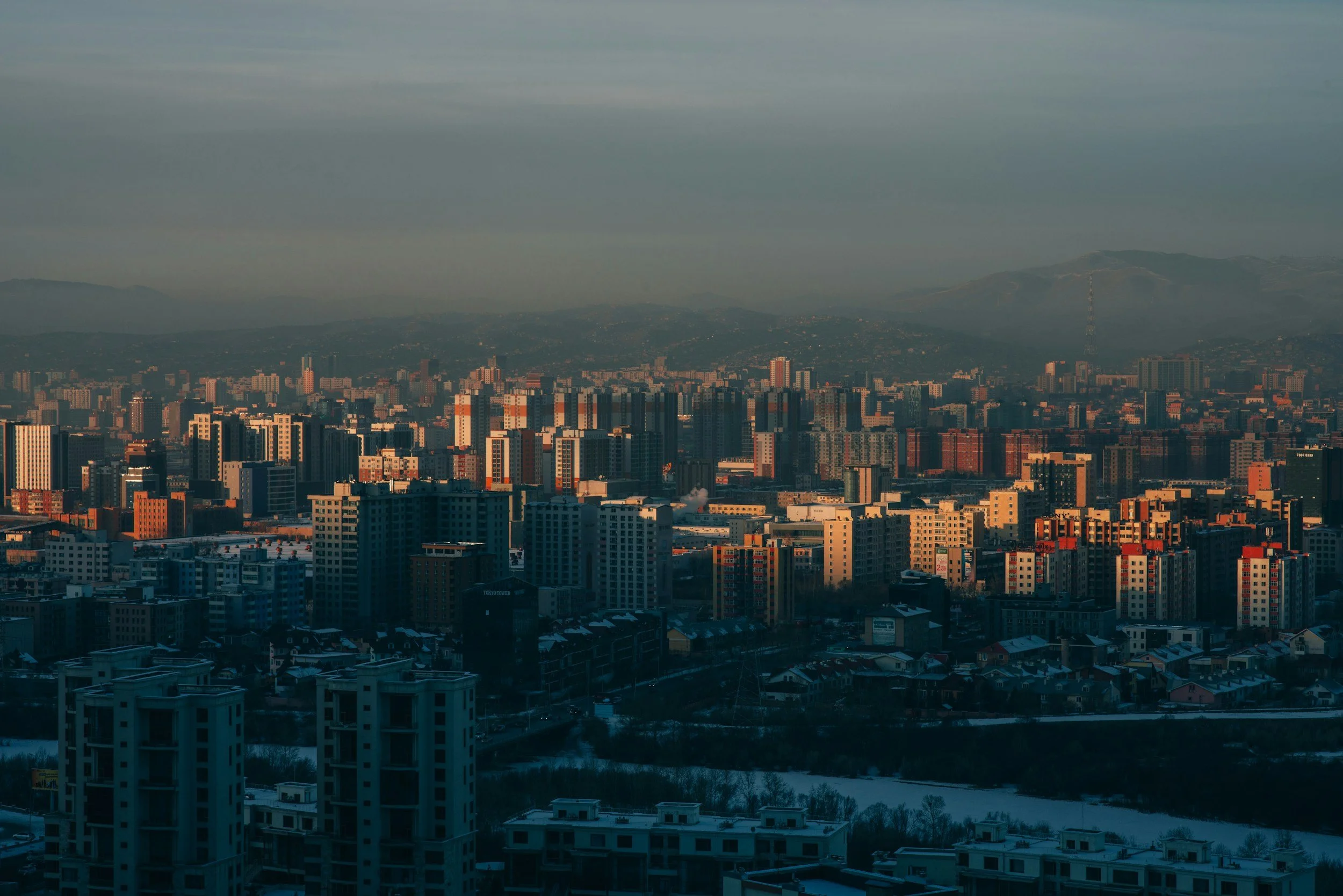 Cityscape with numerous high-rise buildings at sunset, mountains in the background, and snow on the ground.