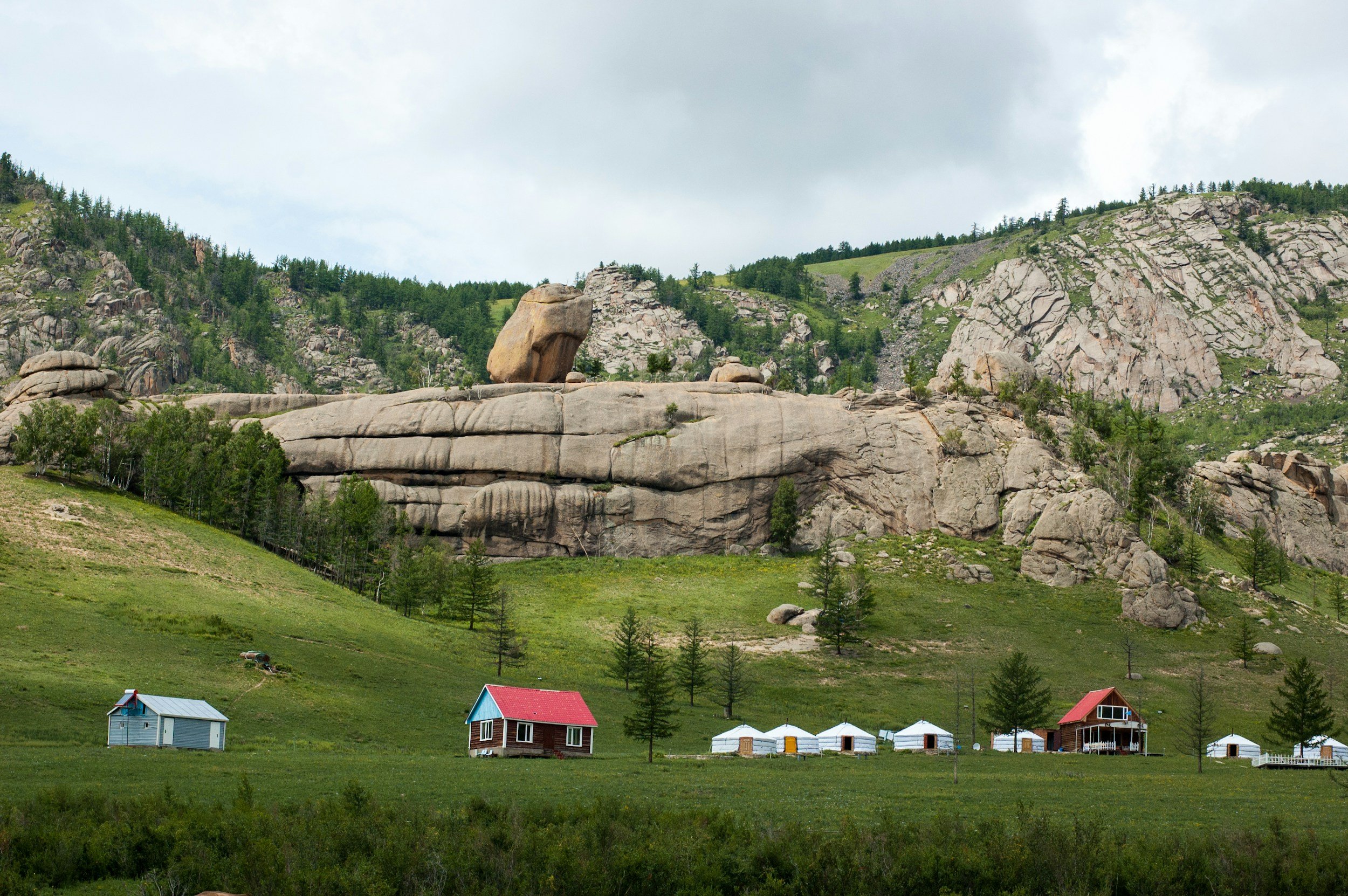 Green valley landscape with small houses, trees, and large rocky mountains in the background under a cloudy sky.