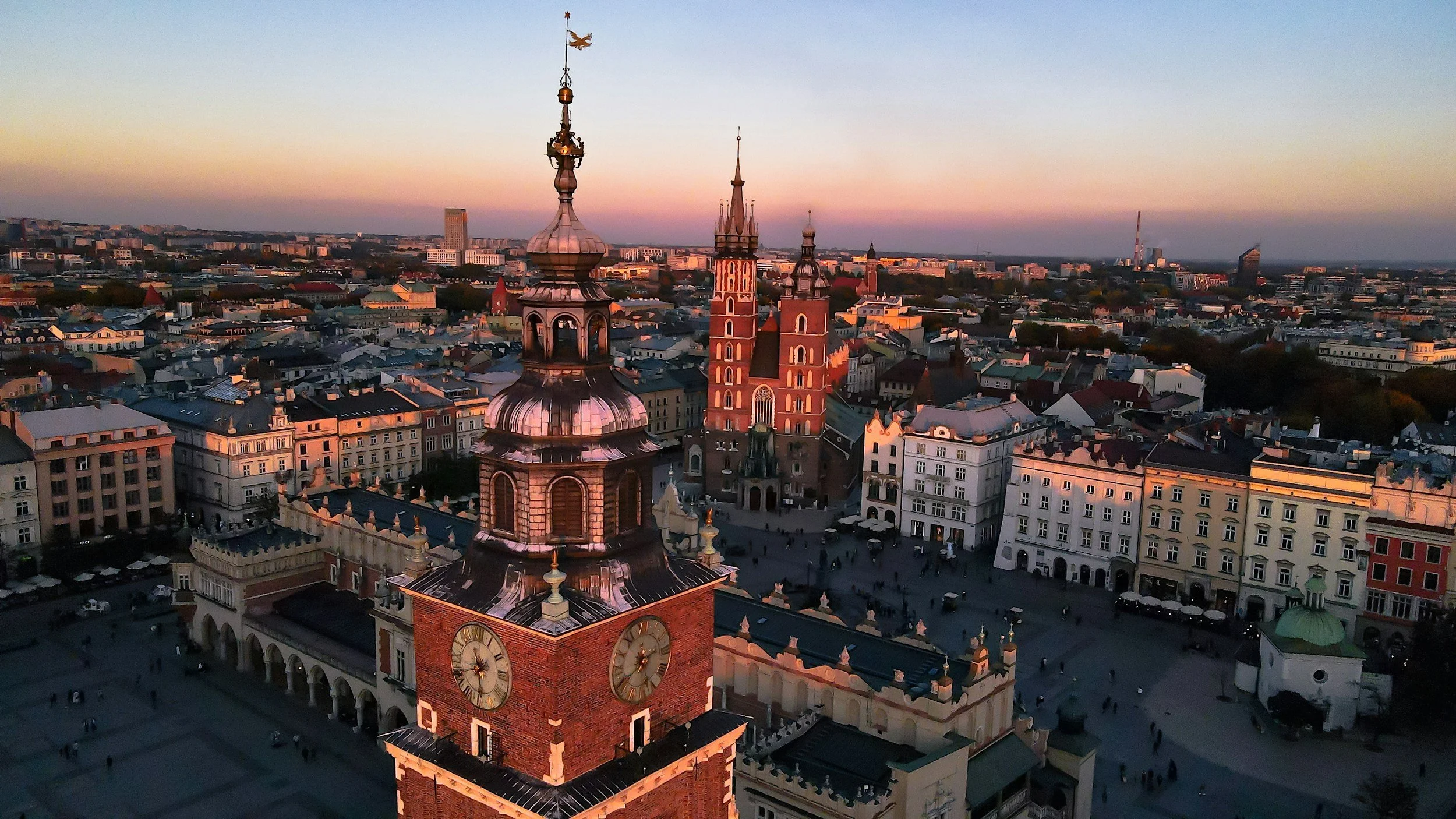 Aerial view of Market Square in Krakow, Poland, at sunset, featuring historic buildings and clocks on a church tower.