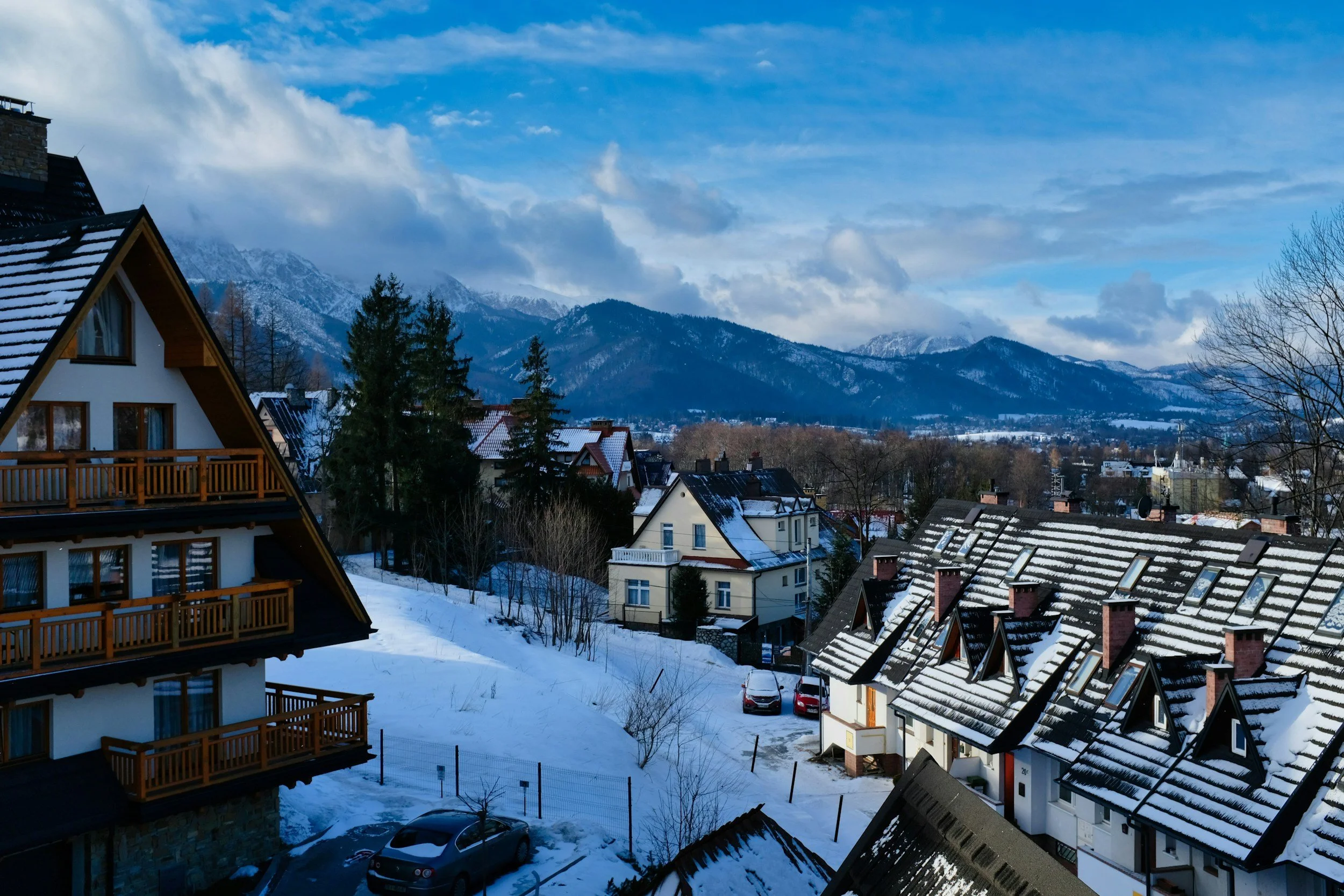 Snow-covered rooftops and houses in a mountain town during daytime with snow-capped mountains and partly cloudy sky in the background