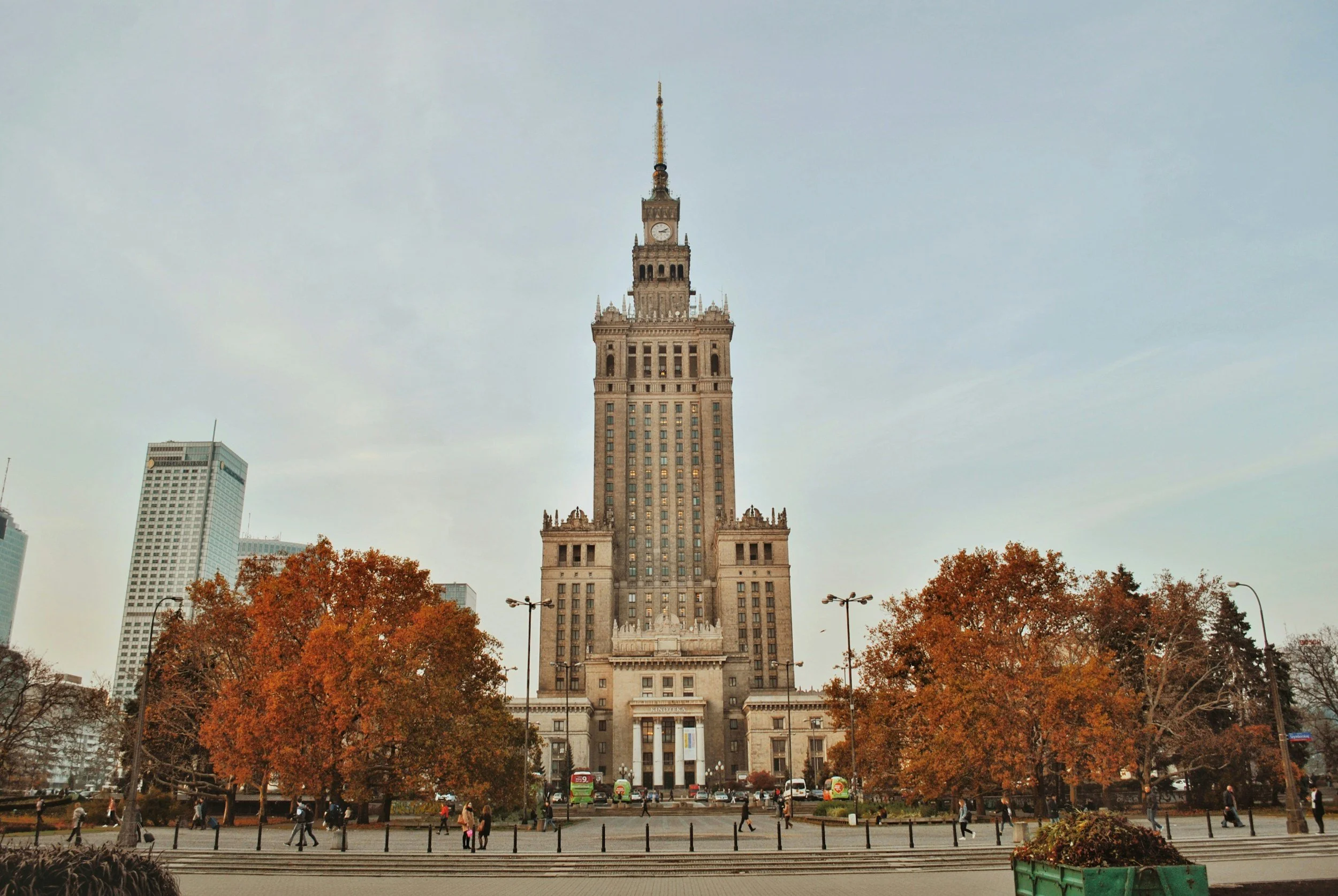 The Palace of Culture and Science in Warsaw, Poland, seen from the front with autumn trees and pedestrians in the foreground.
