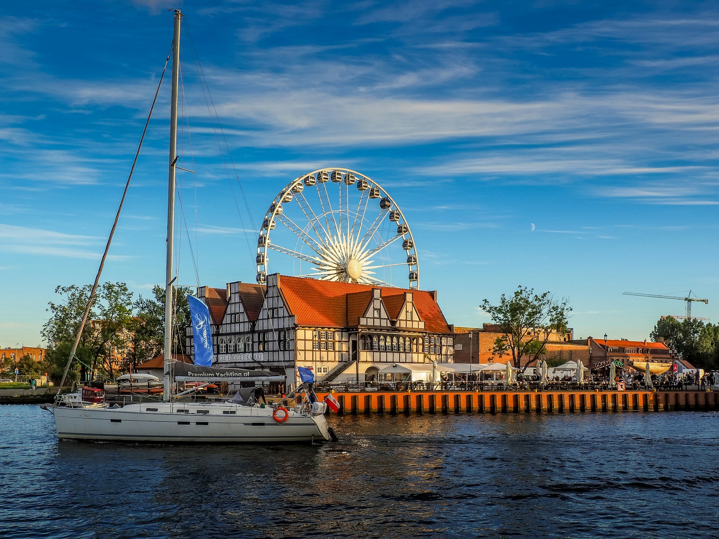 Sailboat docked on water with dockside buildings, a Ferris wheel, and trees in the background under a blue sky with some clouds.