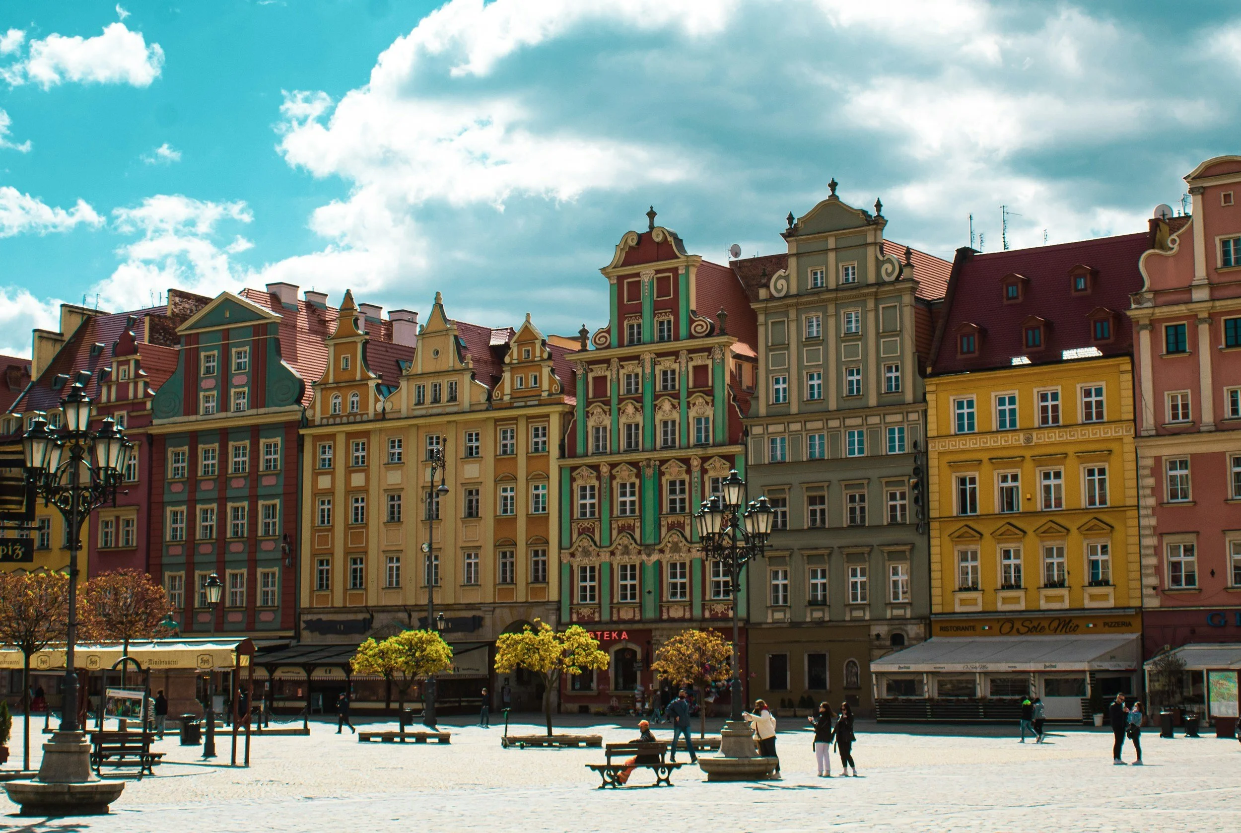 Colorful European buildings with ornate facades along a cobblestone square, sunny sky with clouds, people walking and sitting on benches.