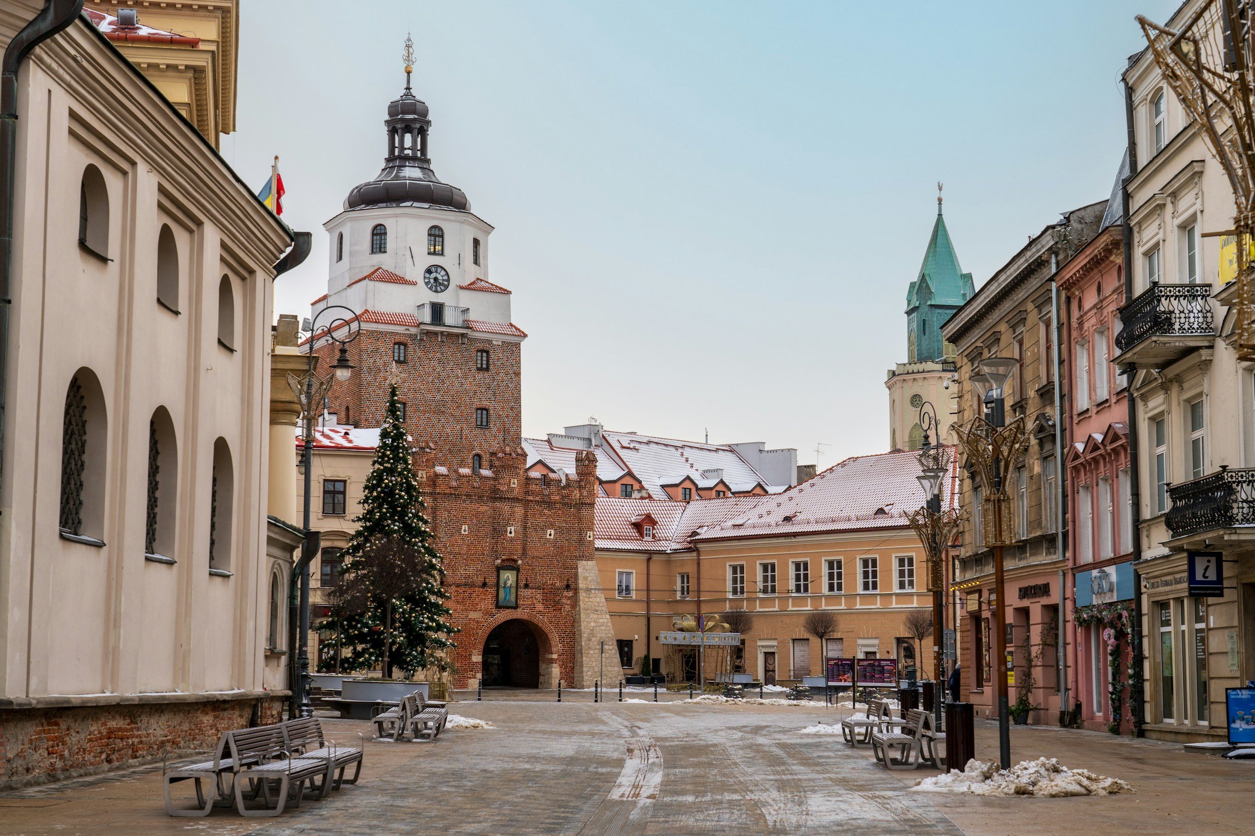 A European city square with a clock tower, colorful buildings, a decorated Christmas tree, and benches on a winter day.