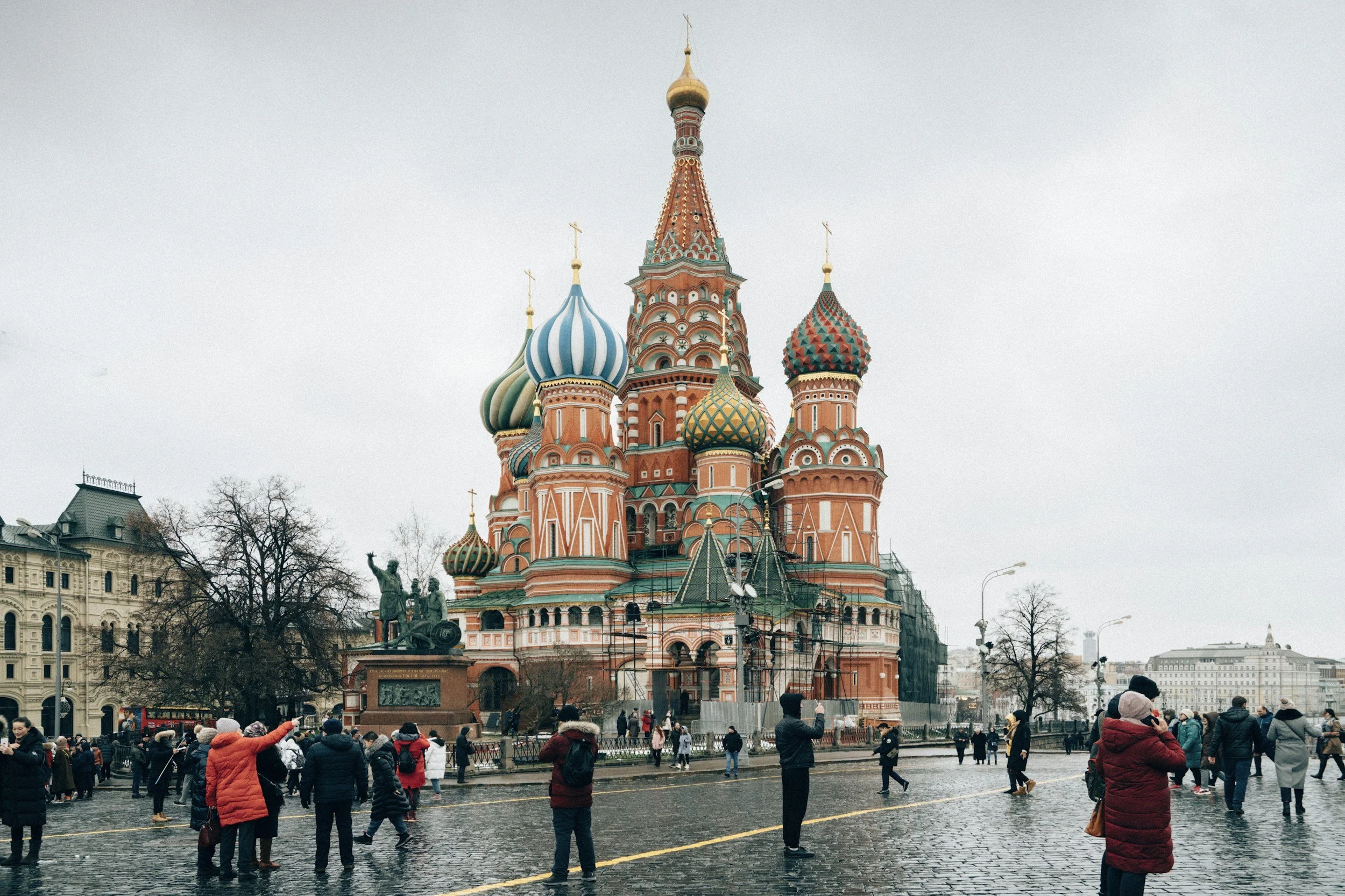 St. Basil's Cathedral in Moscow, Russia, with tourists taking photos and walking on wet cobblestone street on a cloudy day.