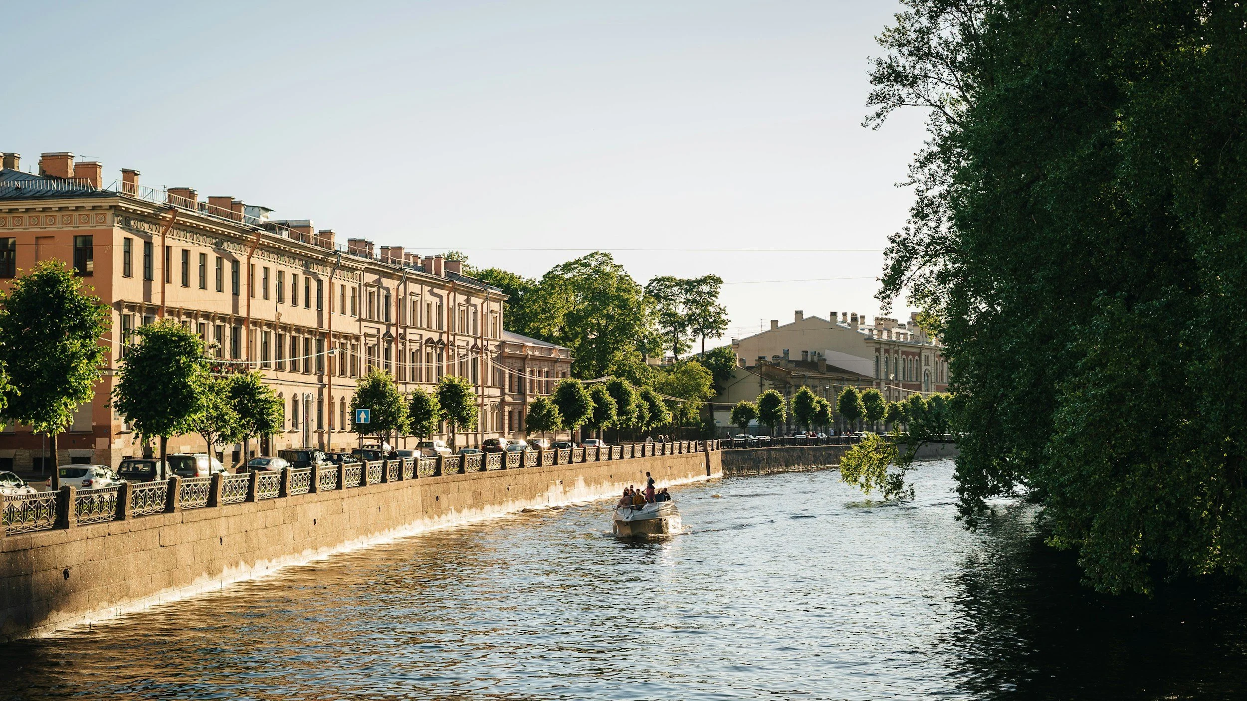 A boat with people sailing on a river beside a city street with parked cars and cobblestone sidewalk, lined with green trees, with historic buildings in the background under a clear sky.