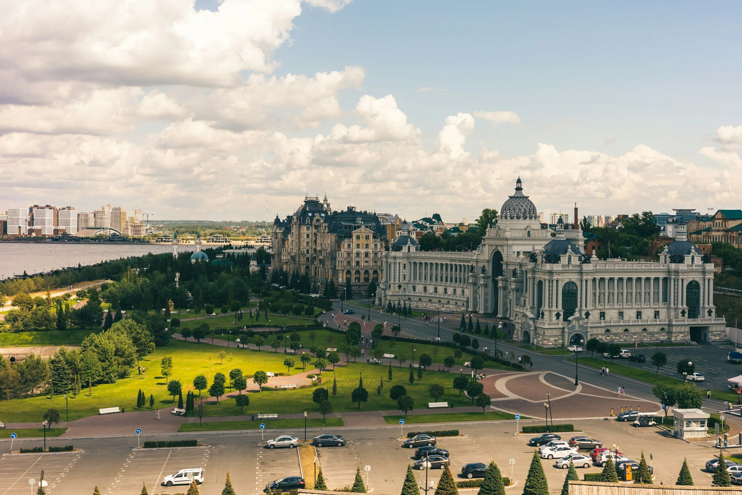 A scenic view of a historic, ornate building with domes and towers, surrounded by a park with green grass, trees, and pathways, and a parking lot in the foreground. City buildings and a river are visible in the background under a partly cloudy sky.