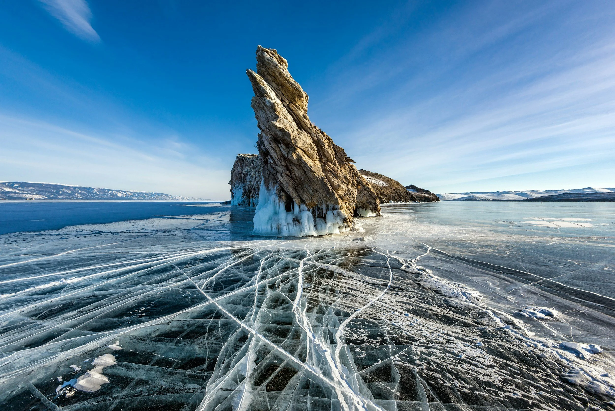 A large rock formation protruding from a frozen lake with cracks in the ice, surrounded by snow-covered hills and a clear blue sky.