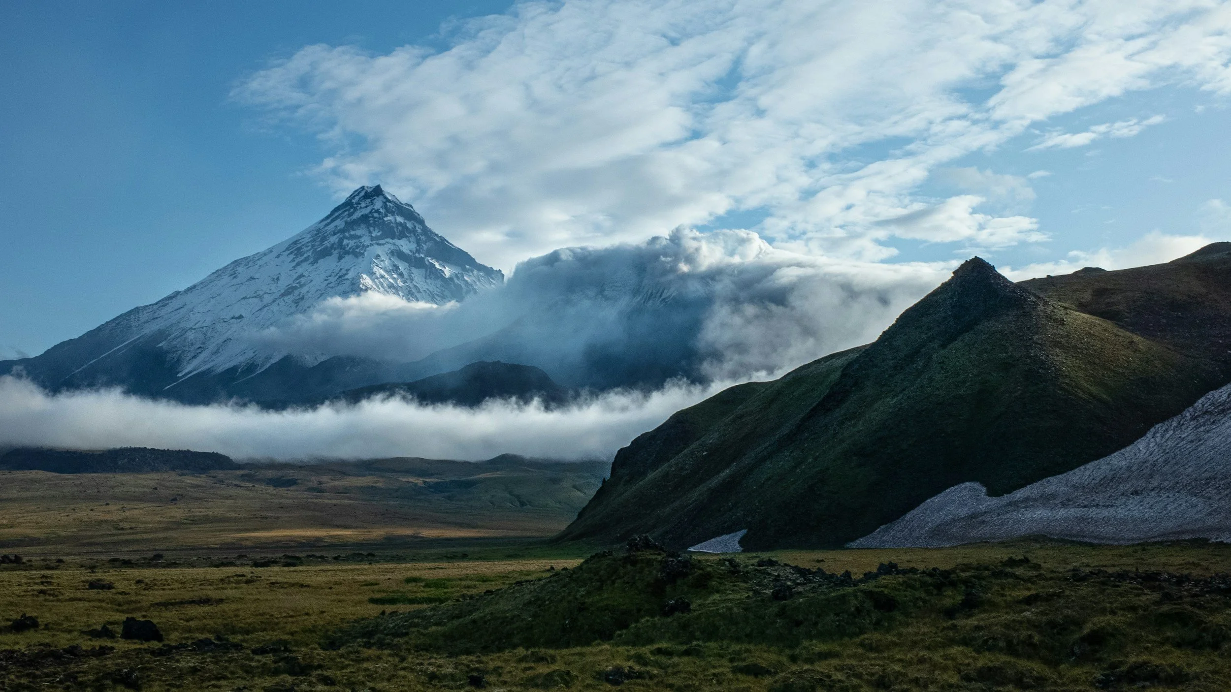 Snow-capped mountain with clouds, green hills, and a grassy plain in the foreground.