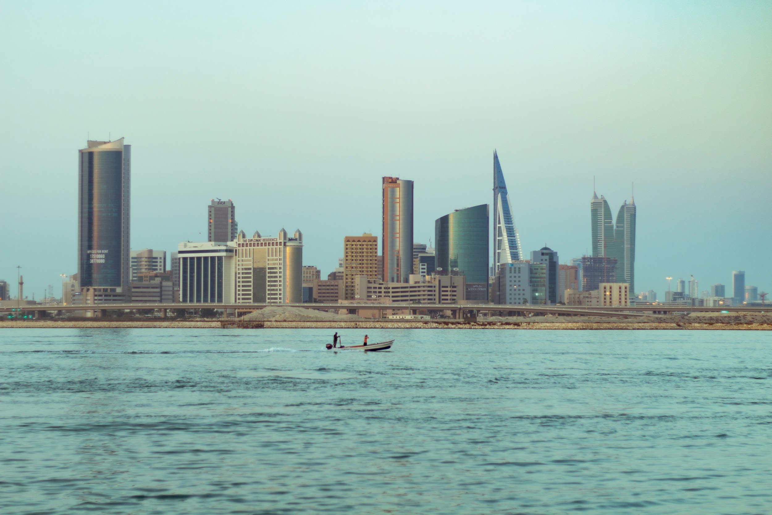 City skyline with tall modern buildings viewed across water, with a small boat and two people in the foreground.