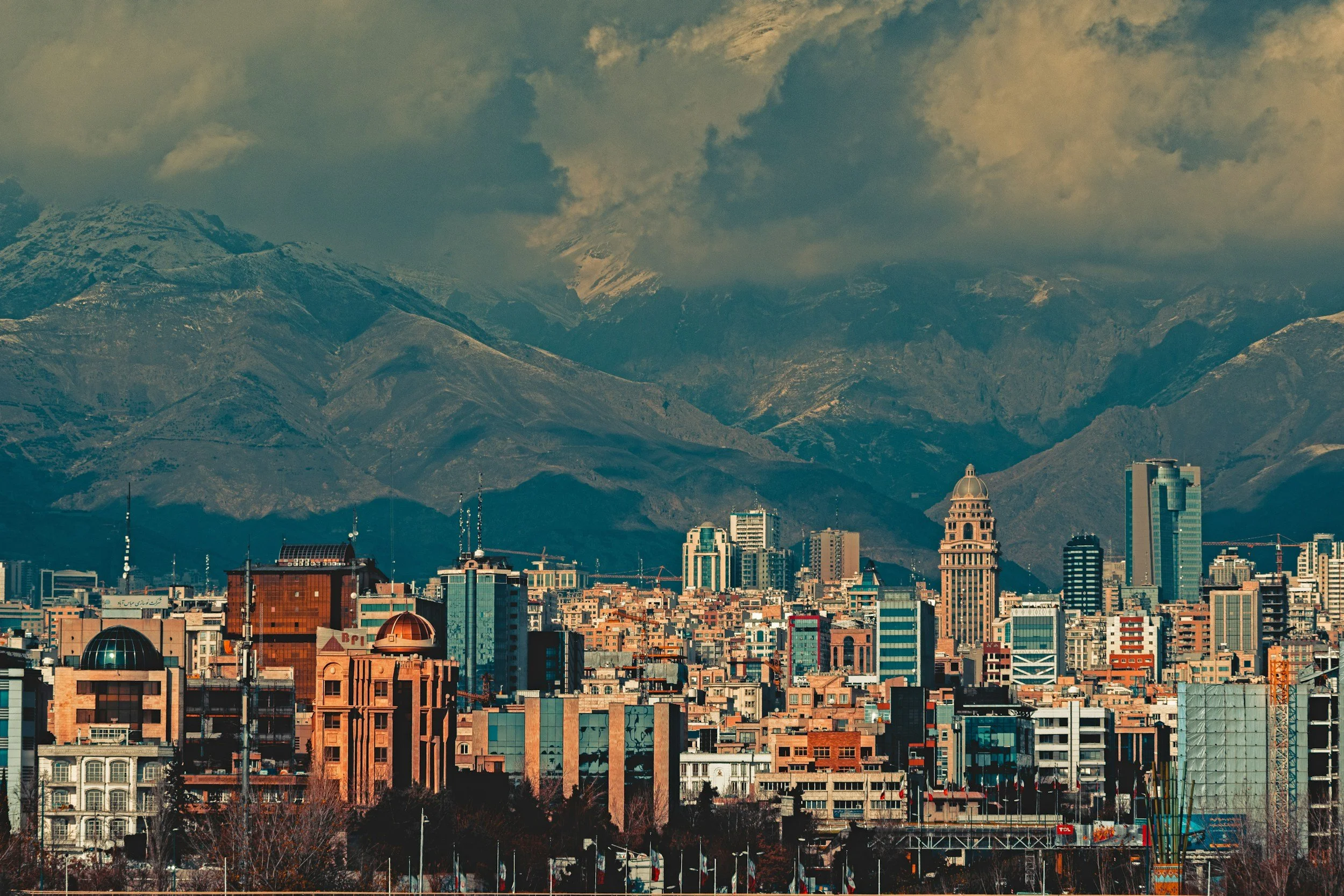 A panoramic view of a city skyline with tall buildings in front of snow-capped mountains under a cloudy sky.