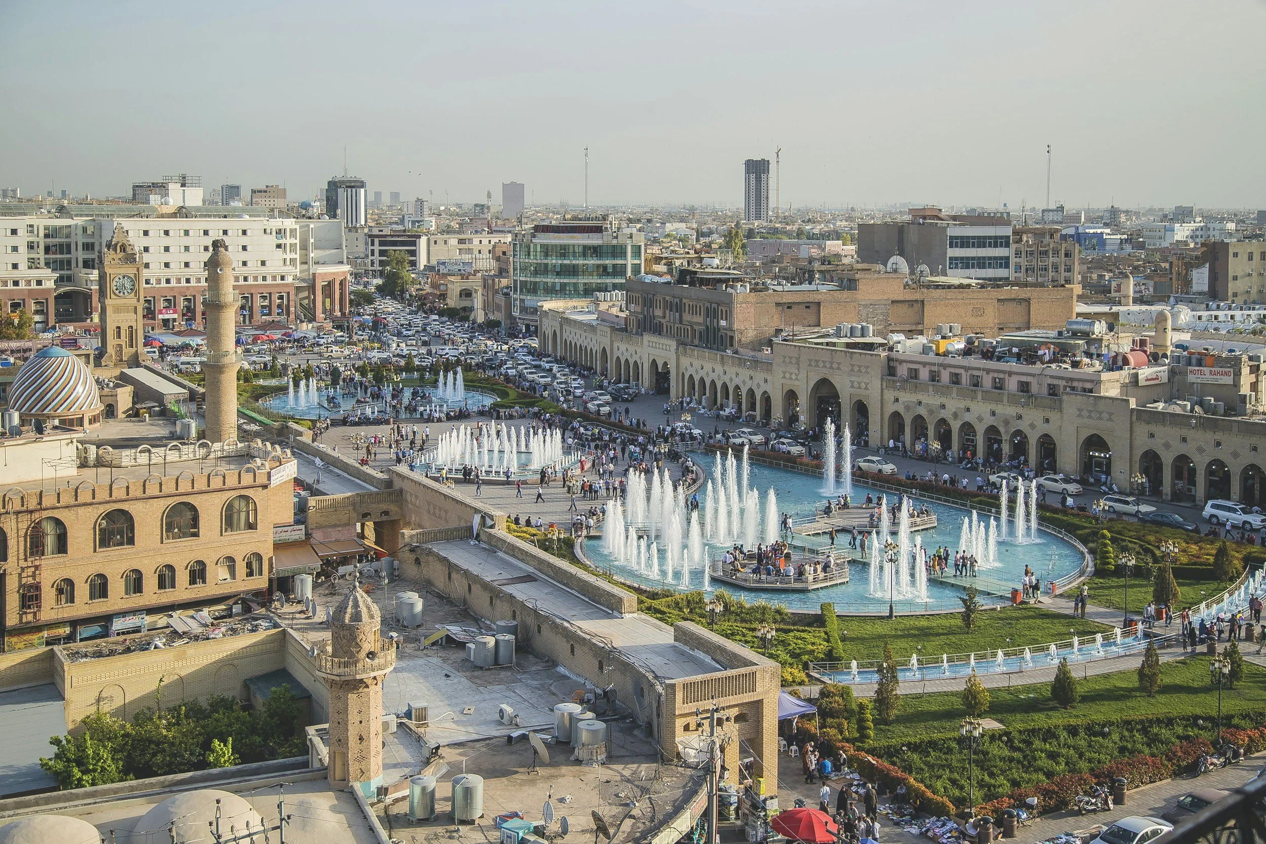 A busy urban park with multiple fountains, surrounded by buildings, with many pedestrians, cars, and a mix of modern and traditional architecture.