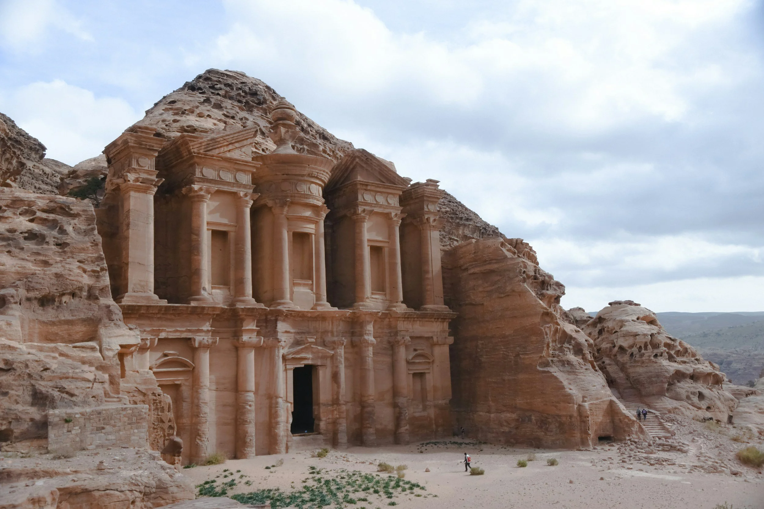 Ancient stone building carved into sandstone cliffs with columns and intricate architecture, set in a desert landscape with a person walking nearby and cloudy sky overhead.