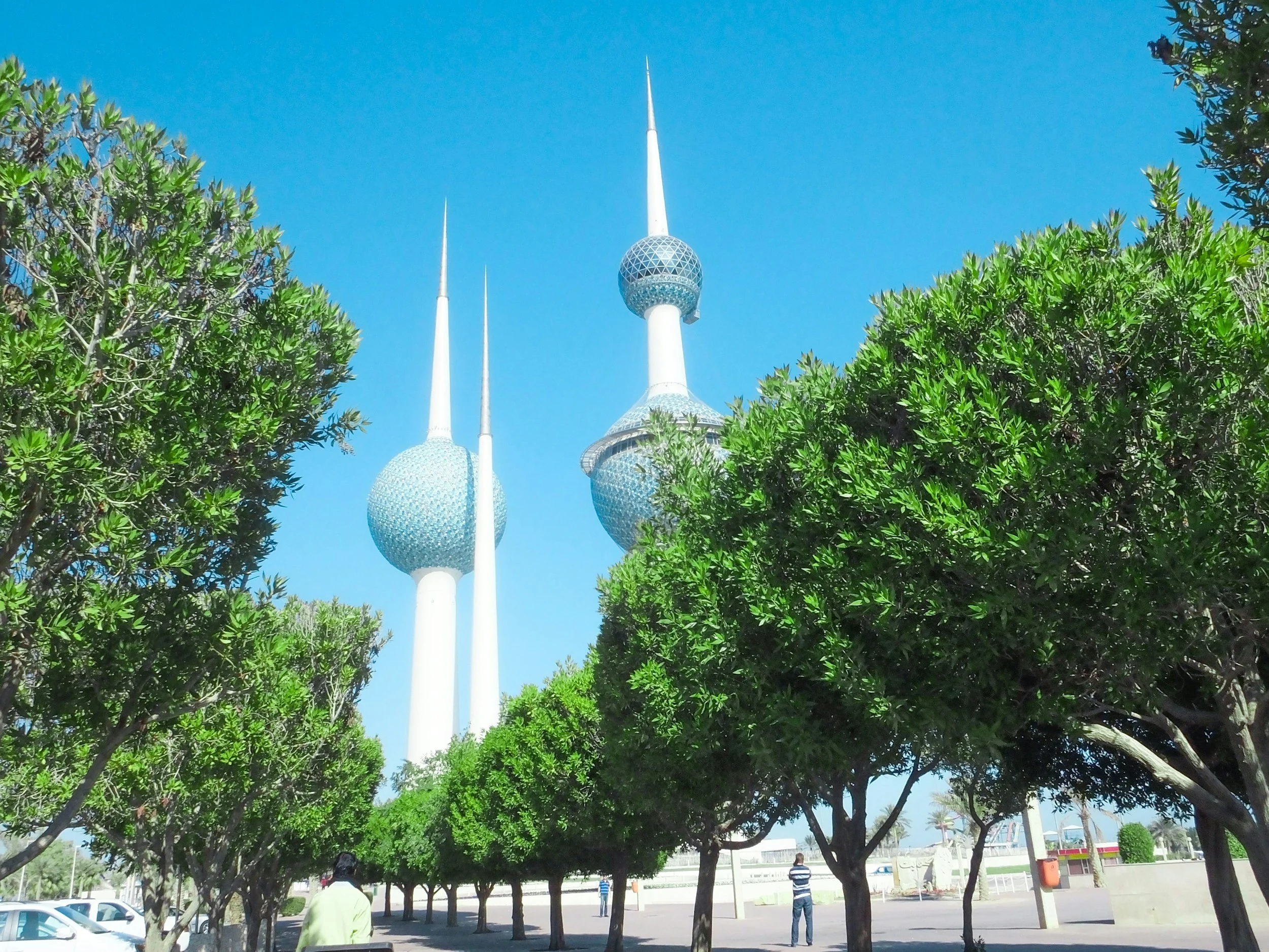 The Kuwait Towers with three tall spires and spherical structures, viewed from below through a row of green trees against a clear blue sky.