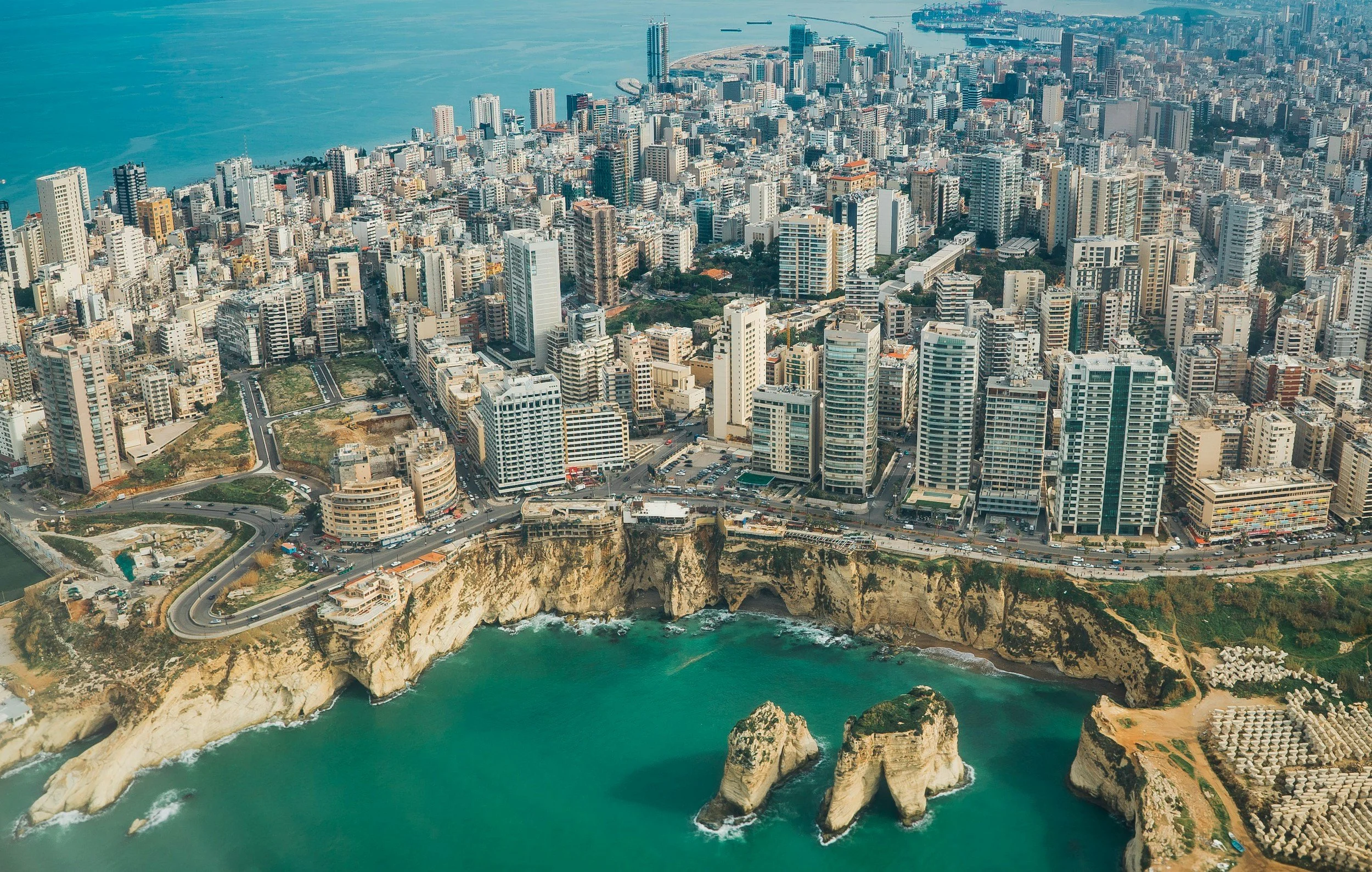 Aerial view of a densely populated city with high-rise buildings along a coastline, featuring cliffs and turquoise water with rock formations.