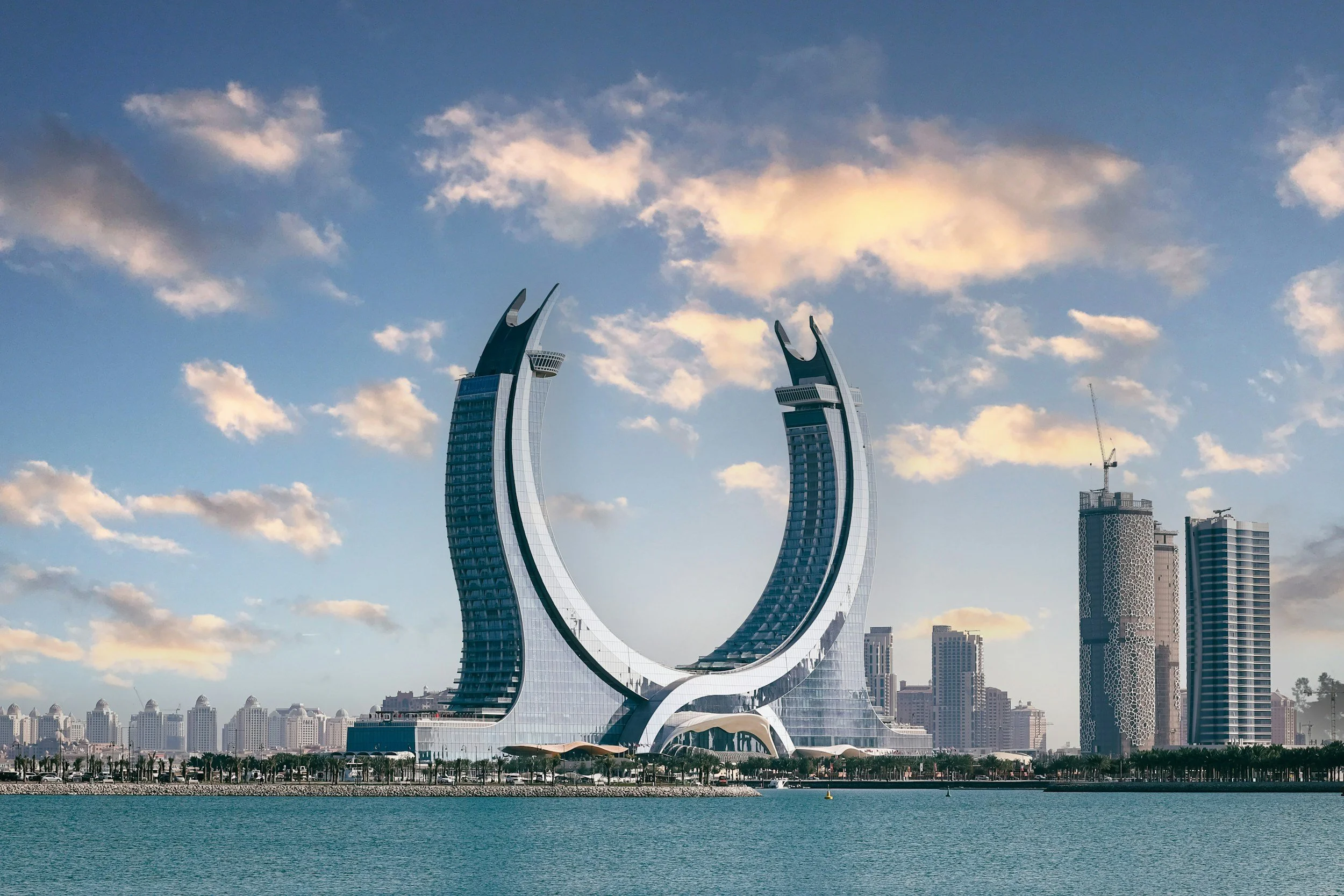 Futuristic twin skyscrapers with curved, crescent shapes, located along a waterfront cityscape, with a cloudy sky overhead. Surrounding buildings and a body of water in the foreground.