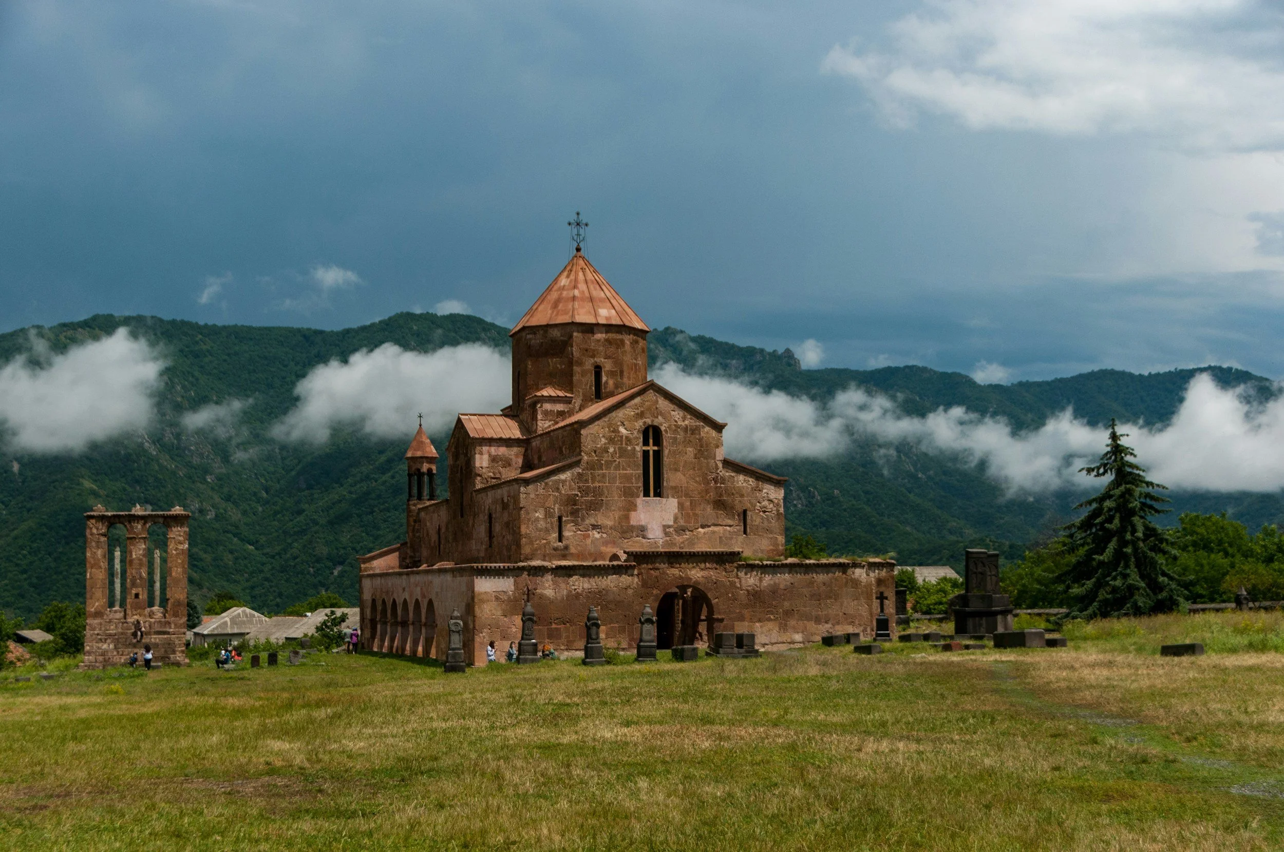 A historic stone church with a central dome and smaller towers, set against green mountains with fog and a cloudy sky.