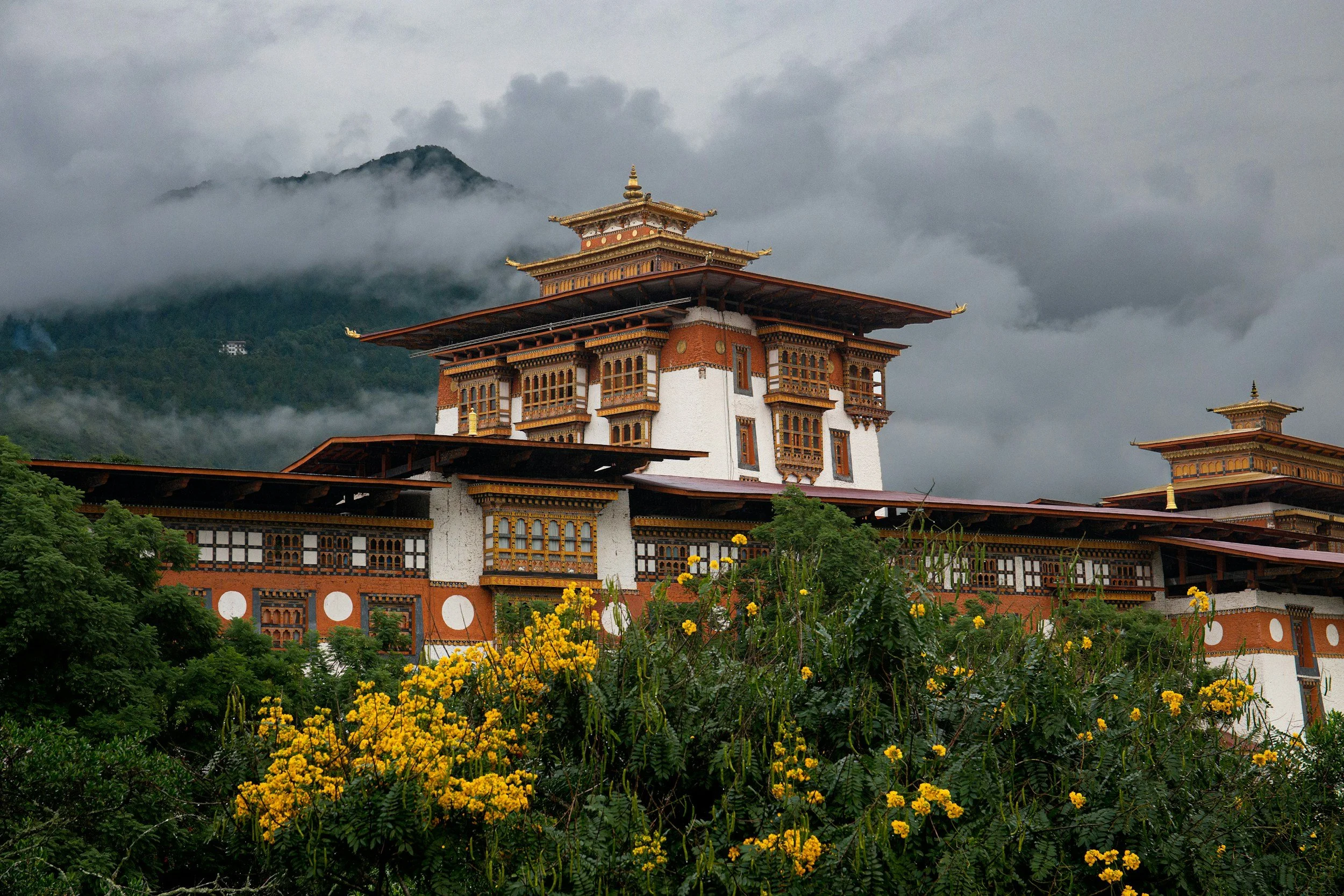 A traditional Bhutanese fortress or temple with intricate wooden details, set against a cloudy mountain background and surrounded by lush greenery with yellow flowers in the foreground.