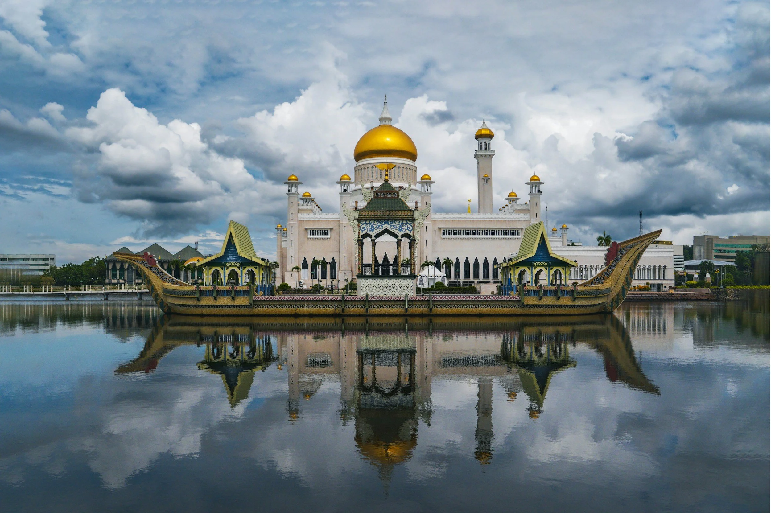 The image shows the Sultan Omar Ali Saifuddien Mosque located in Brunei, reflected perfectly in the water in front of it, under a cloudy sky.