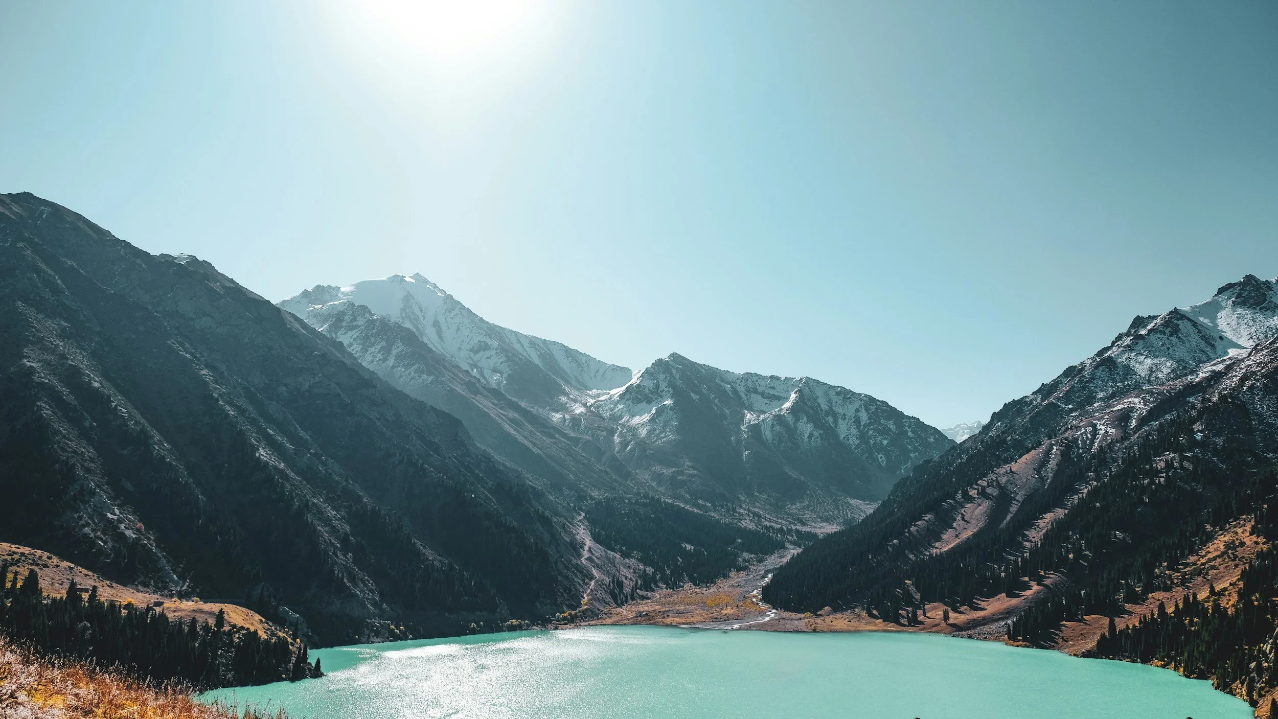 Mountains surrounding a turquoise lake with snow-capped peaks and forested slopes under a clear sky.