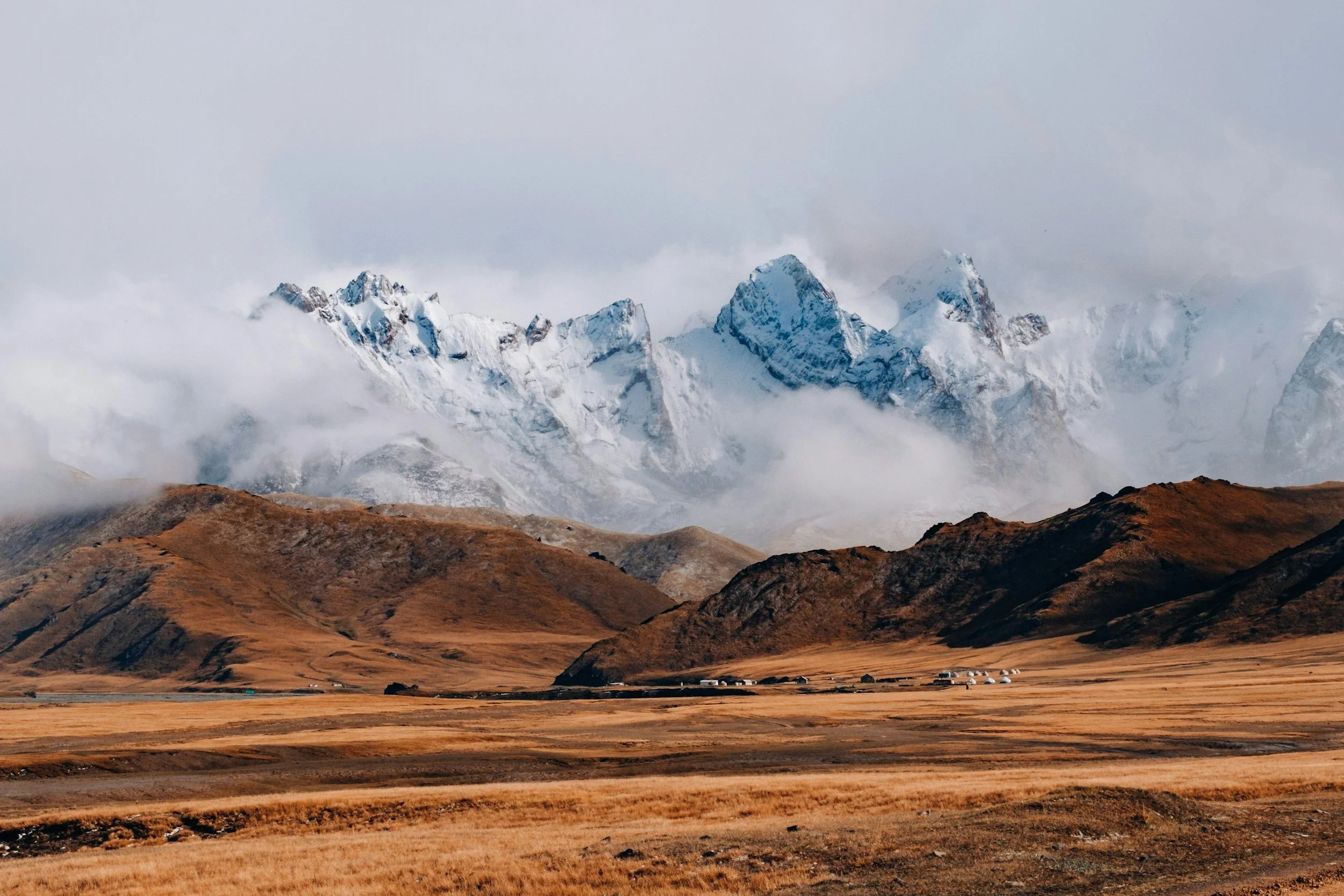 A landscape of snow-capped mountains with mist and clouds, alongside rolling brown grassy hills and a few small houses or buildings in the foreground.