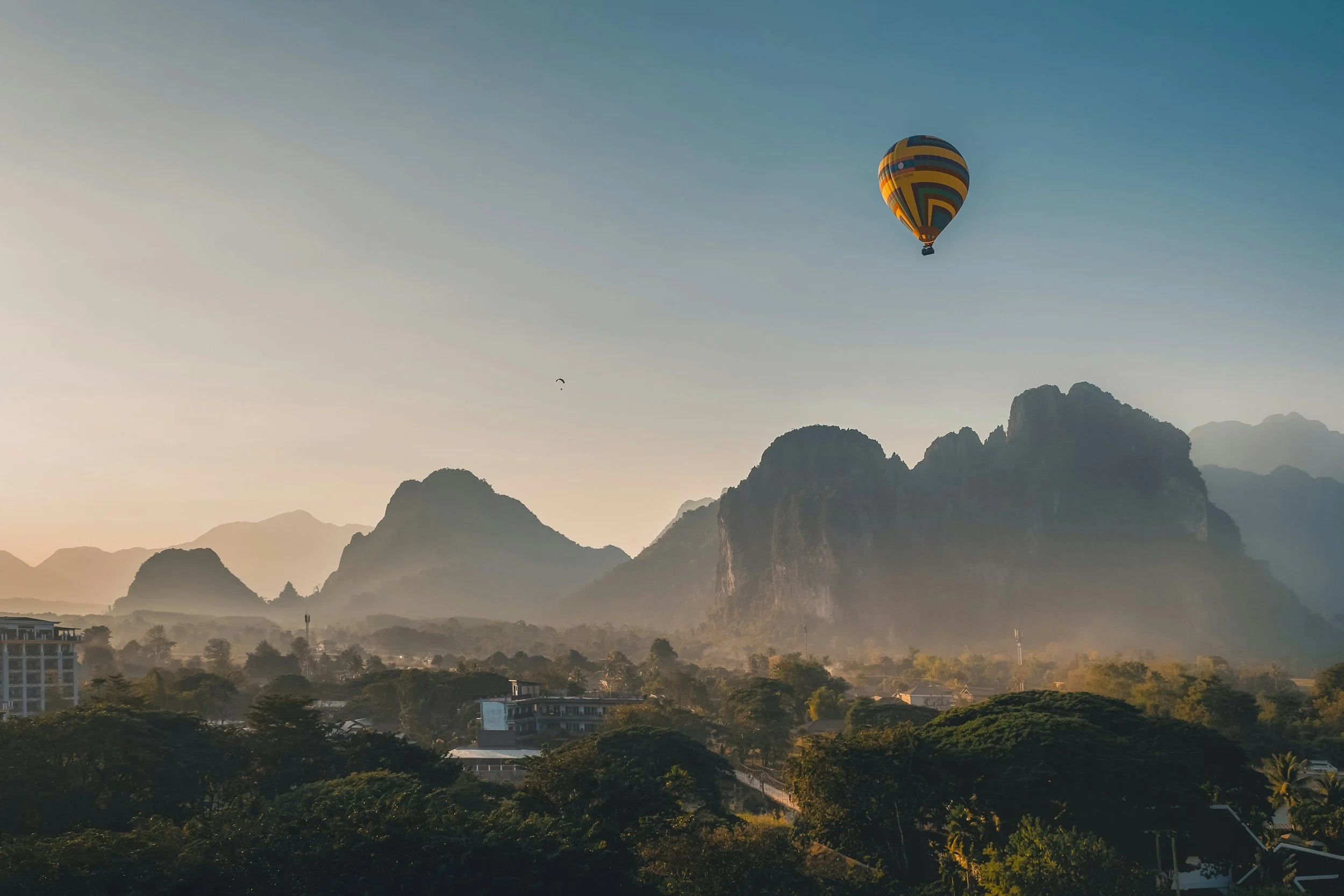 A hot air balloon with colorful geometric patterns flying over lush green mountains at sunrise.