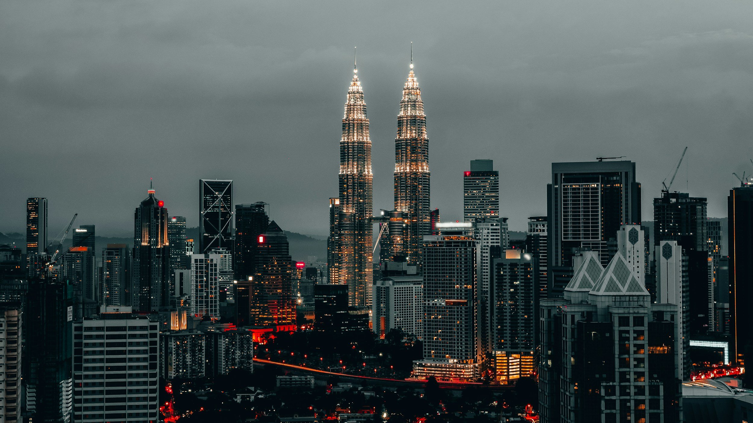 Nighttime cityscape of Kuala Lumpur featuring the illuminated Petronas Twin Towers against dark cloudy sky