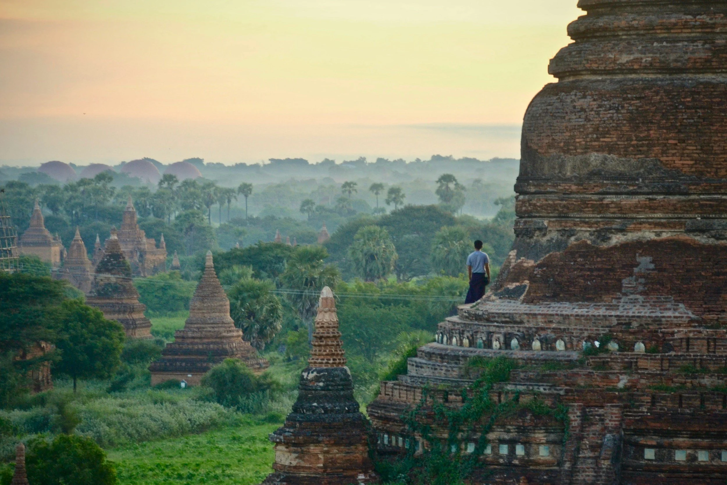 A person standing on a large brick temple in an ancient temple complex surrounded by greenery and smaller temples at sunset in Bagan, Myanmar.