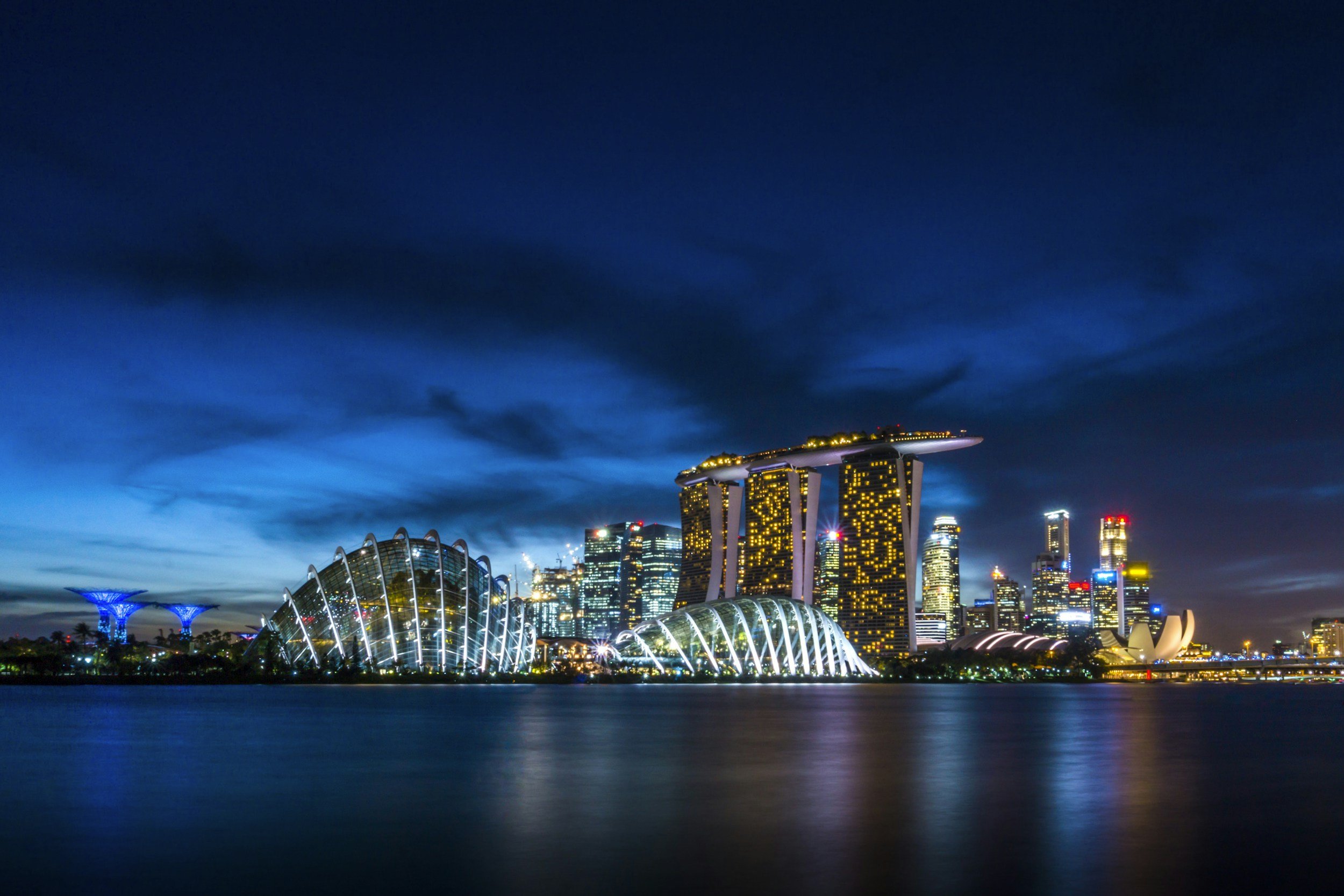 Night view of Singapore skyline with Marina Bay Sands hotel, flower-shaped ArtScience Museum, and city skyscrapers, reflected on water.