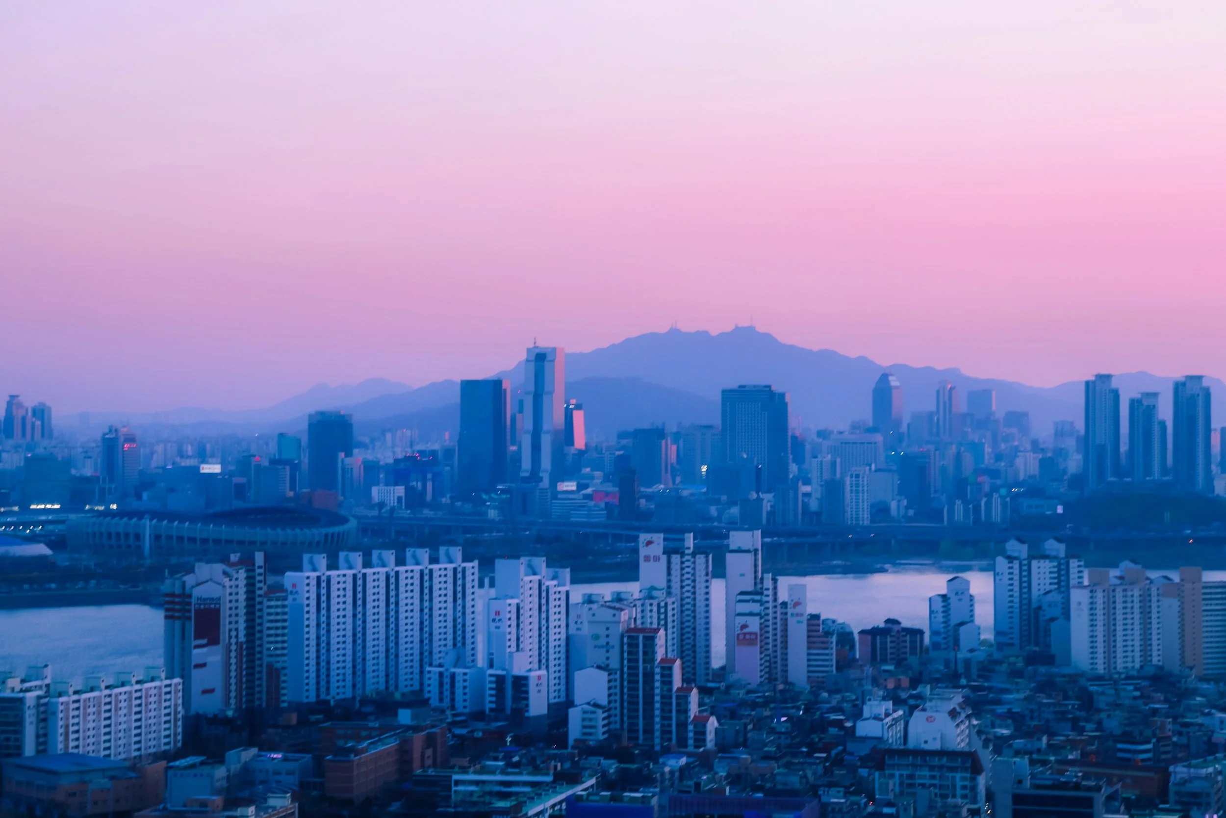 City skyline at dusk with tall buildings, a river in the foreground, and mountains in the background under a colorful sky.