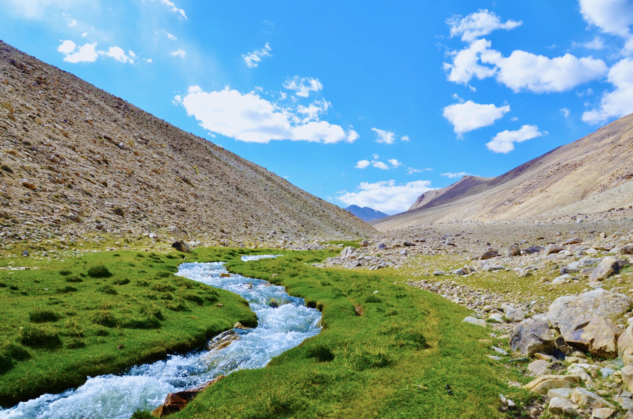 A mountain valley with a small winding river flowing through lush green grass, flanked by rocky hills and a bright blue sky with white clouds.