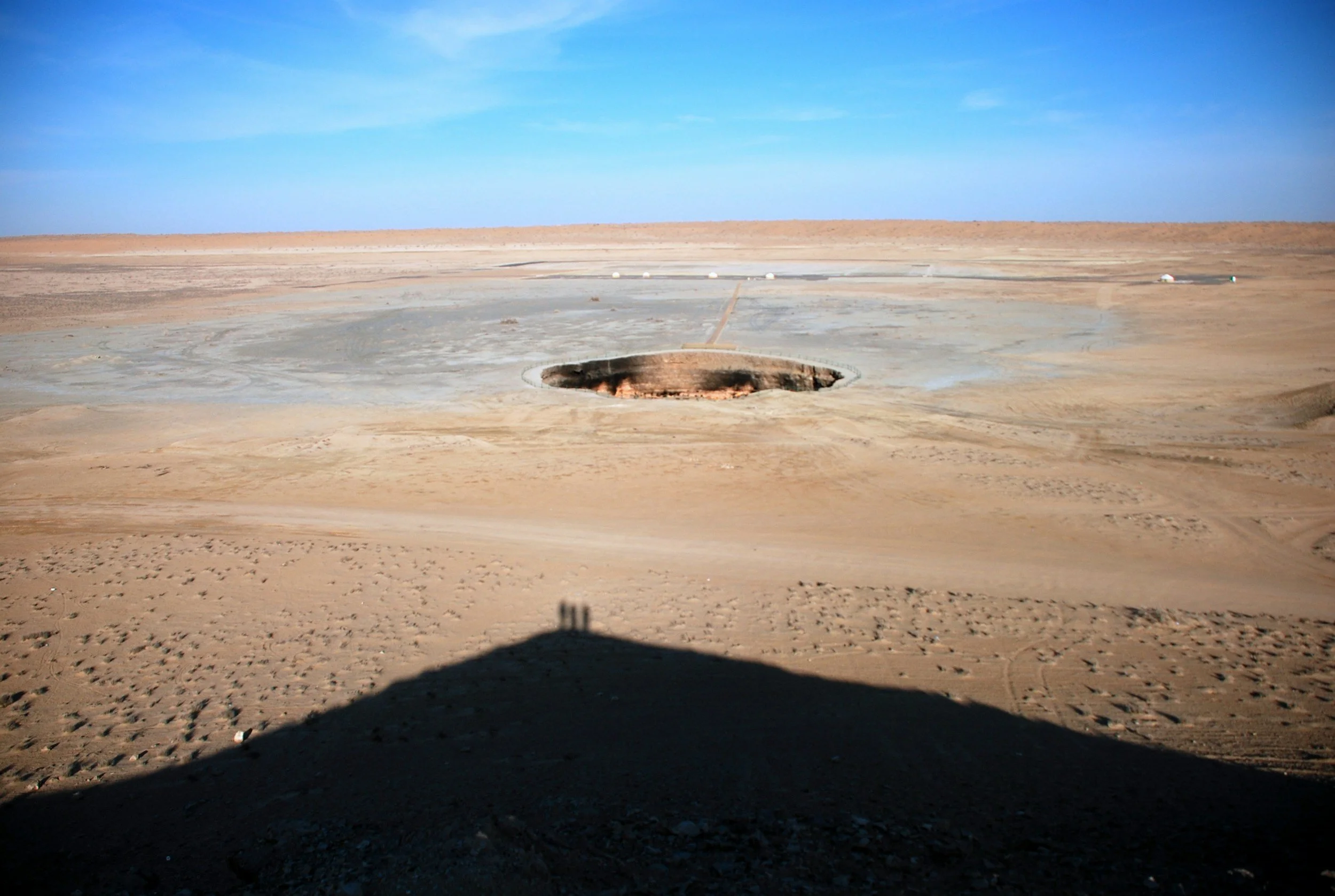 Large sinkhole in desert landscape with clear blue sky, sparse terrain, and a shadow in the foreground.