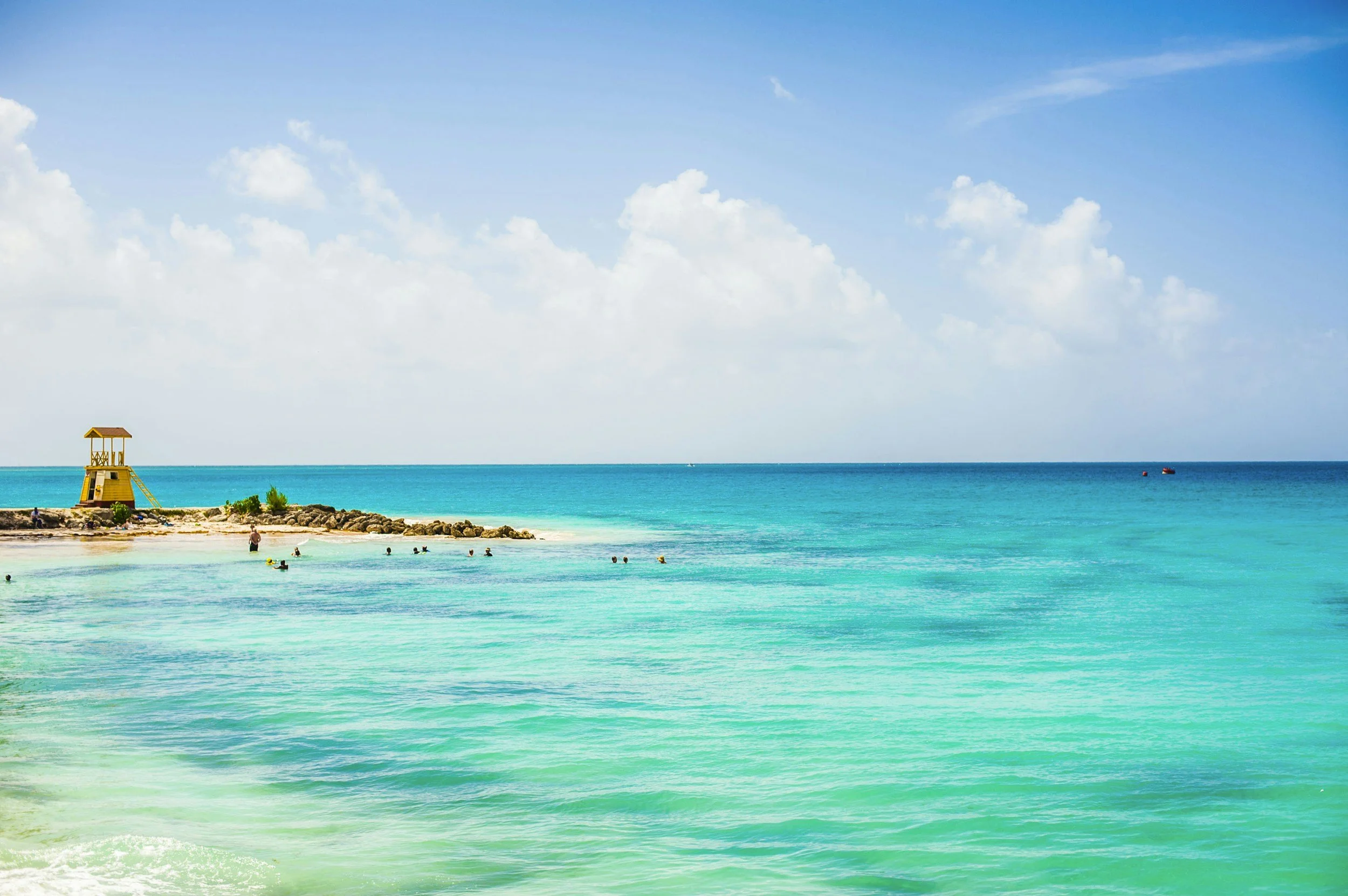 A tropical beach scene with turquoise water, a sandy shore, a small yellow lifeguard tower, and people swimming in the ocean. There are clouds in the blue sky.